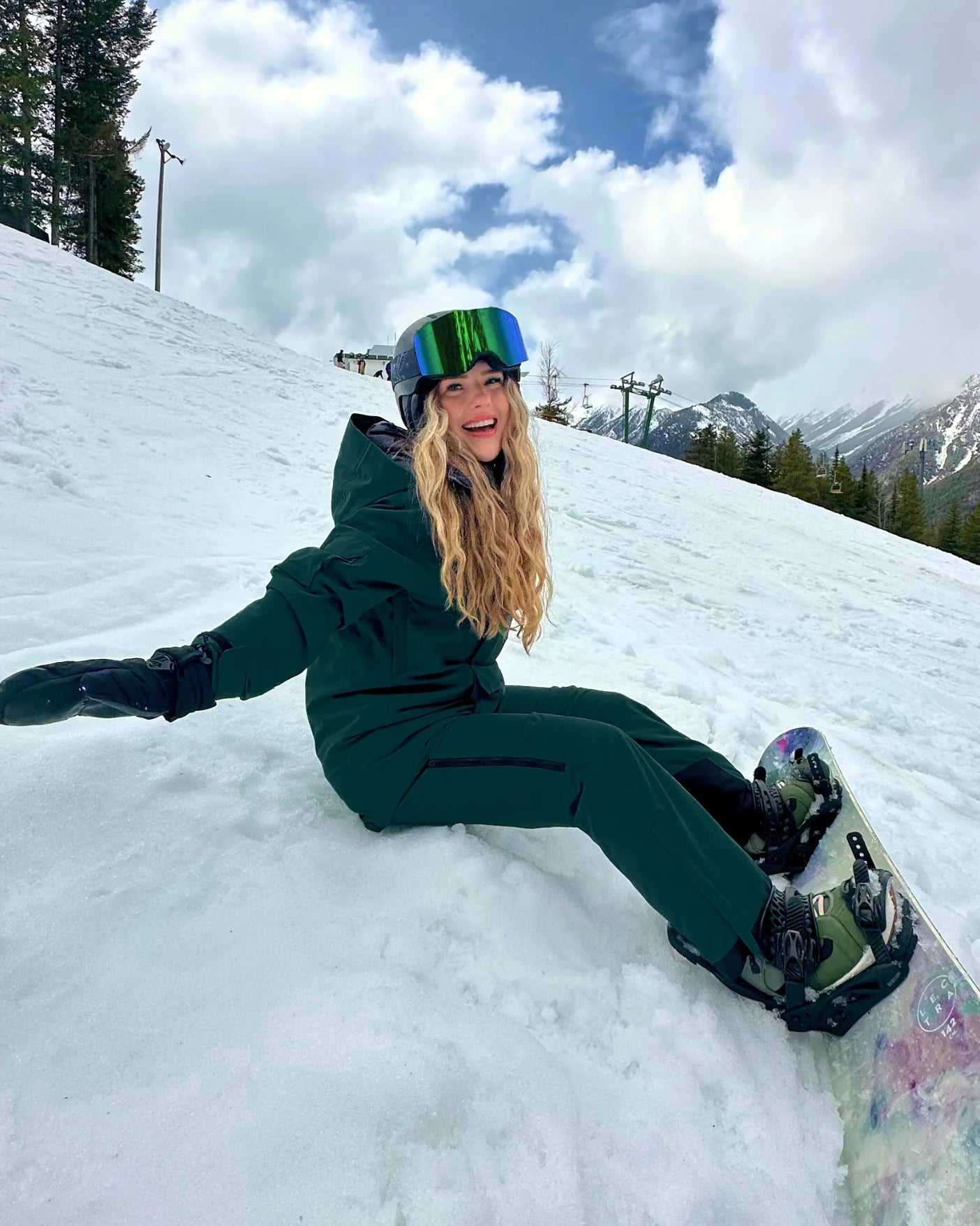 Woman wearing Nexarina emerald green bib snow pants, seated on snow with snowboard in a mountain setting.