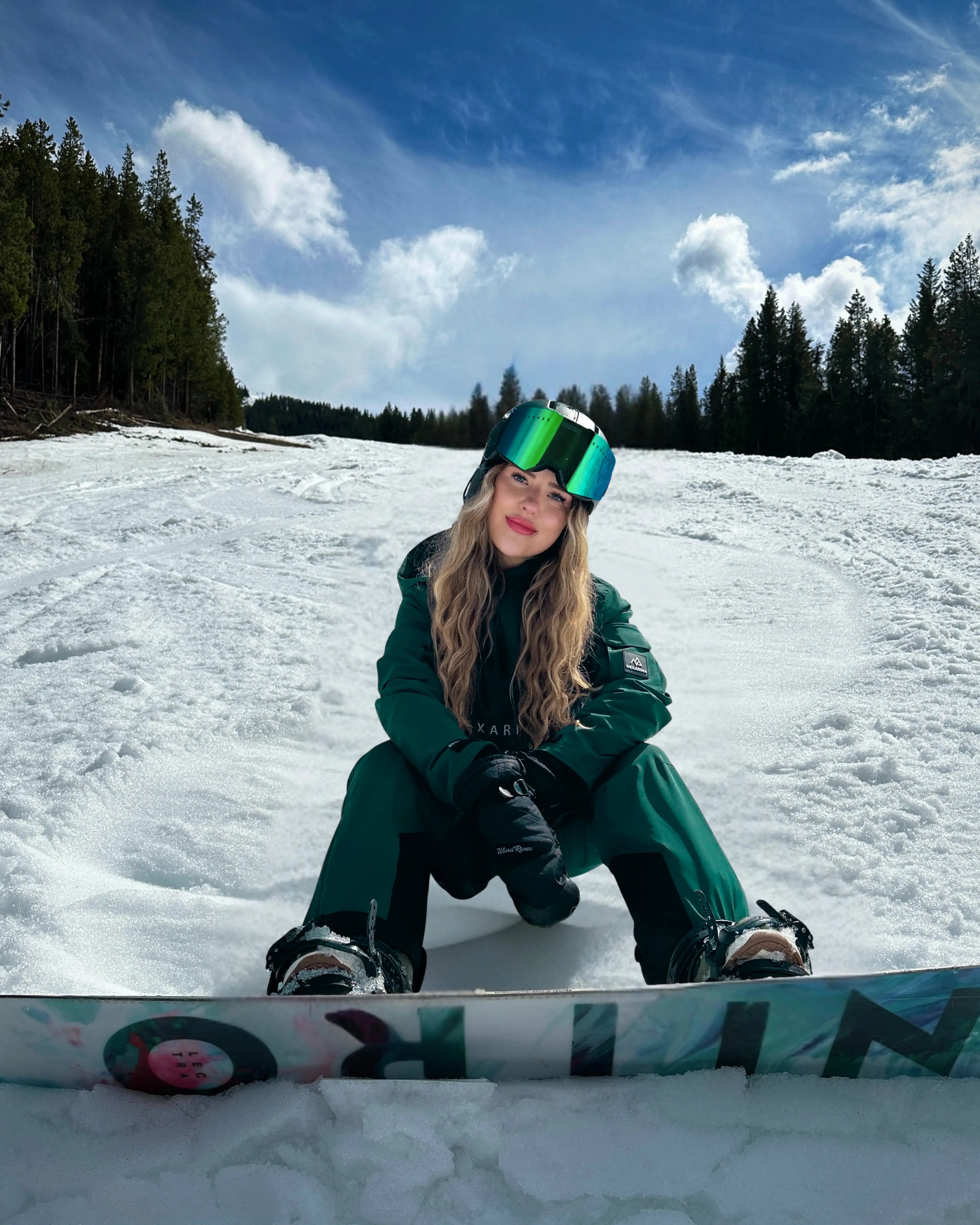 Female snowboarder wearing Nexarina green bib pants in a mountain snow setting.
