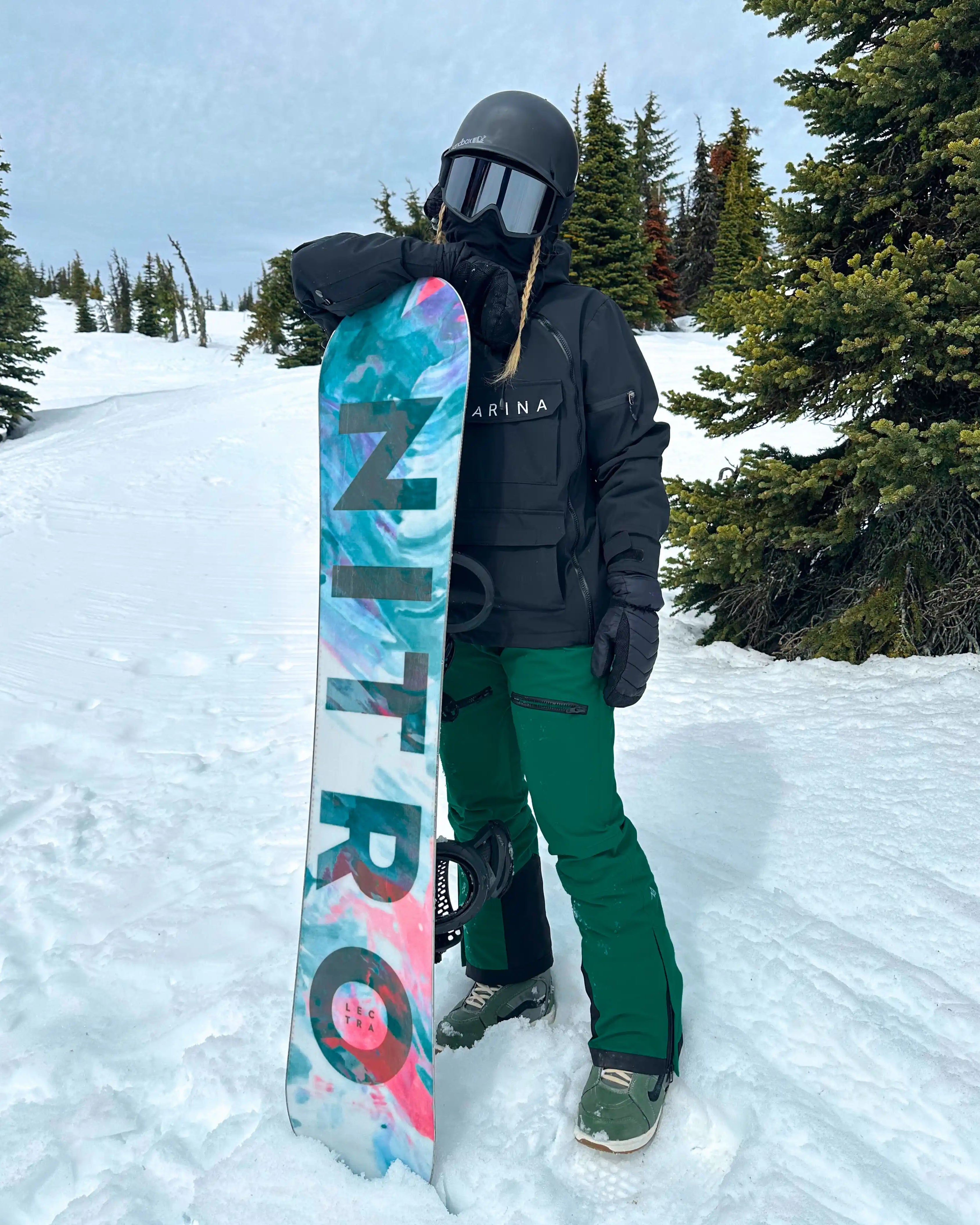 Woman standing in snowy mountain terrain holding a colorful Nitro snowboard, wearing a black Nexarina jacket and emerald green snow pants, helmet and goggles on, representing women’s technical outerwear.