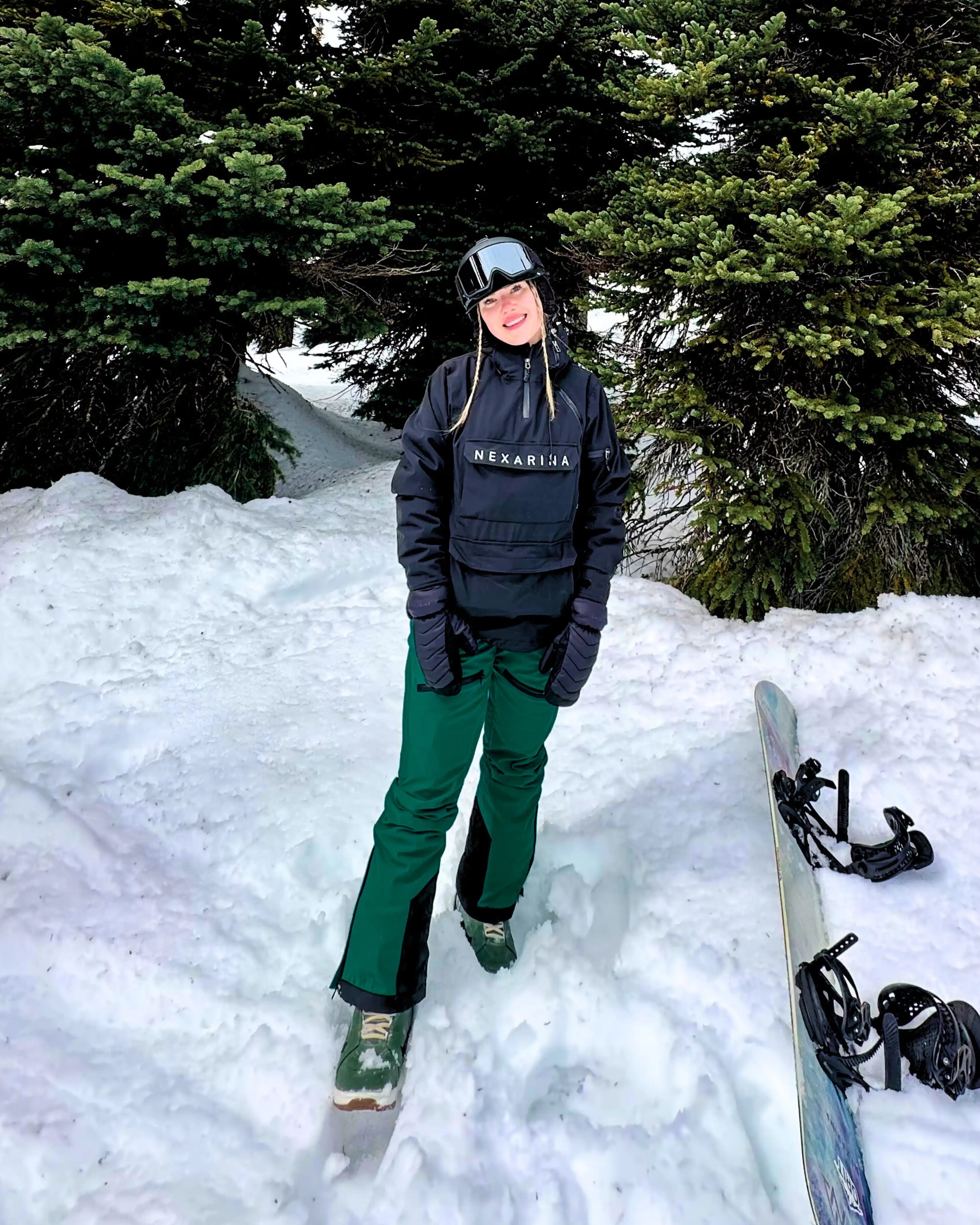 Woman smiling in snowy forest setting, dressed in a black Nexarina jacket and emerald green snow pants, helmet and goggles off, with a snowboard resting in the snow beside her.
