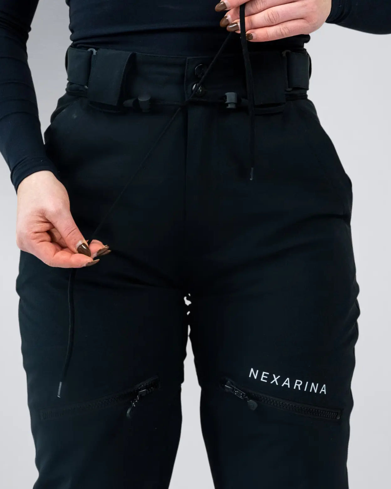 Front view of Nexarina Avani Snow Pant in Classic Black, featuring zippered thigh pockets and green boots.