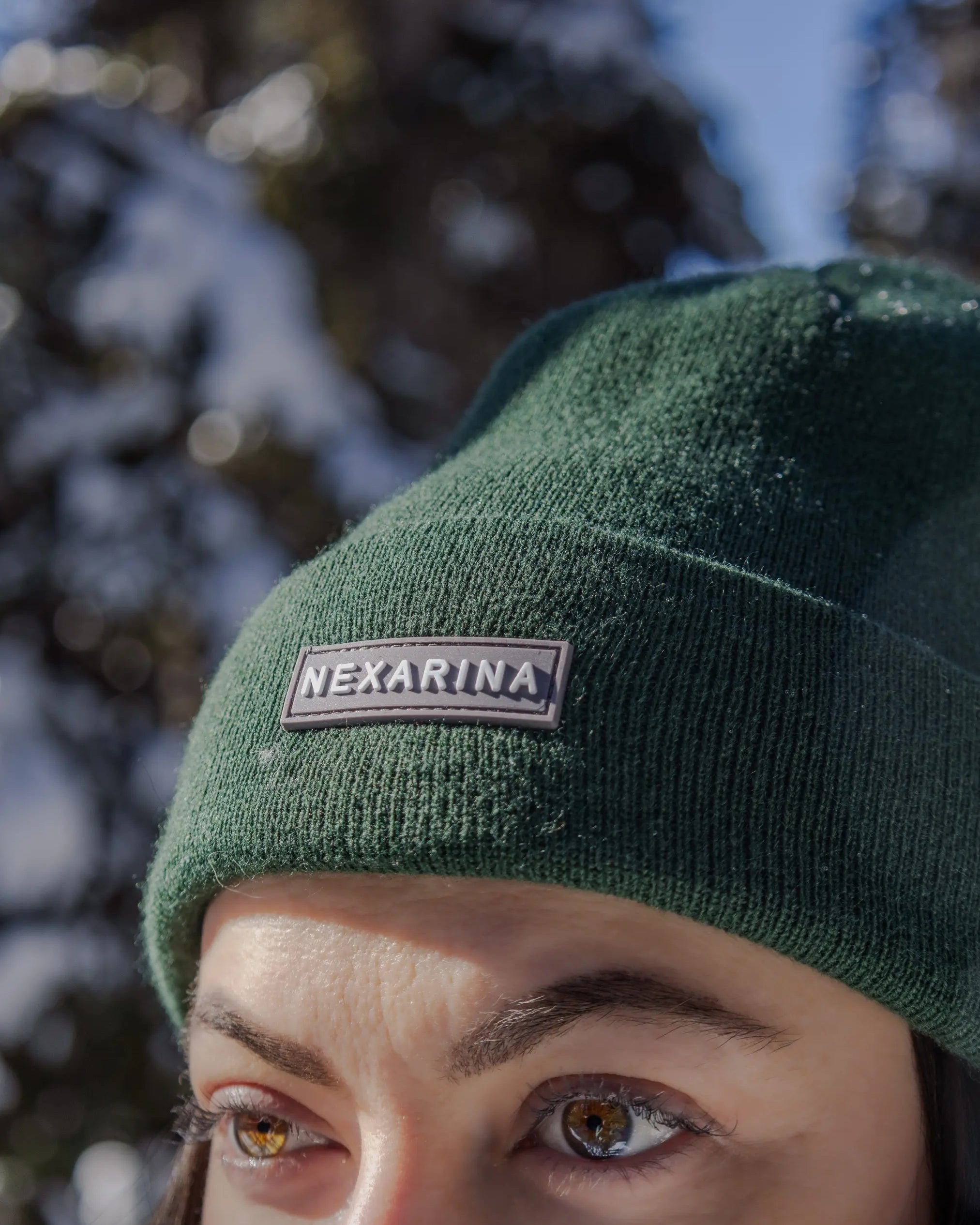 Close-up of a forest green Nexarina beanie with logo patch, worn outdoors with snow-covered trees in the background.