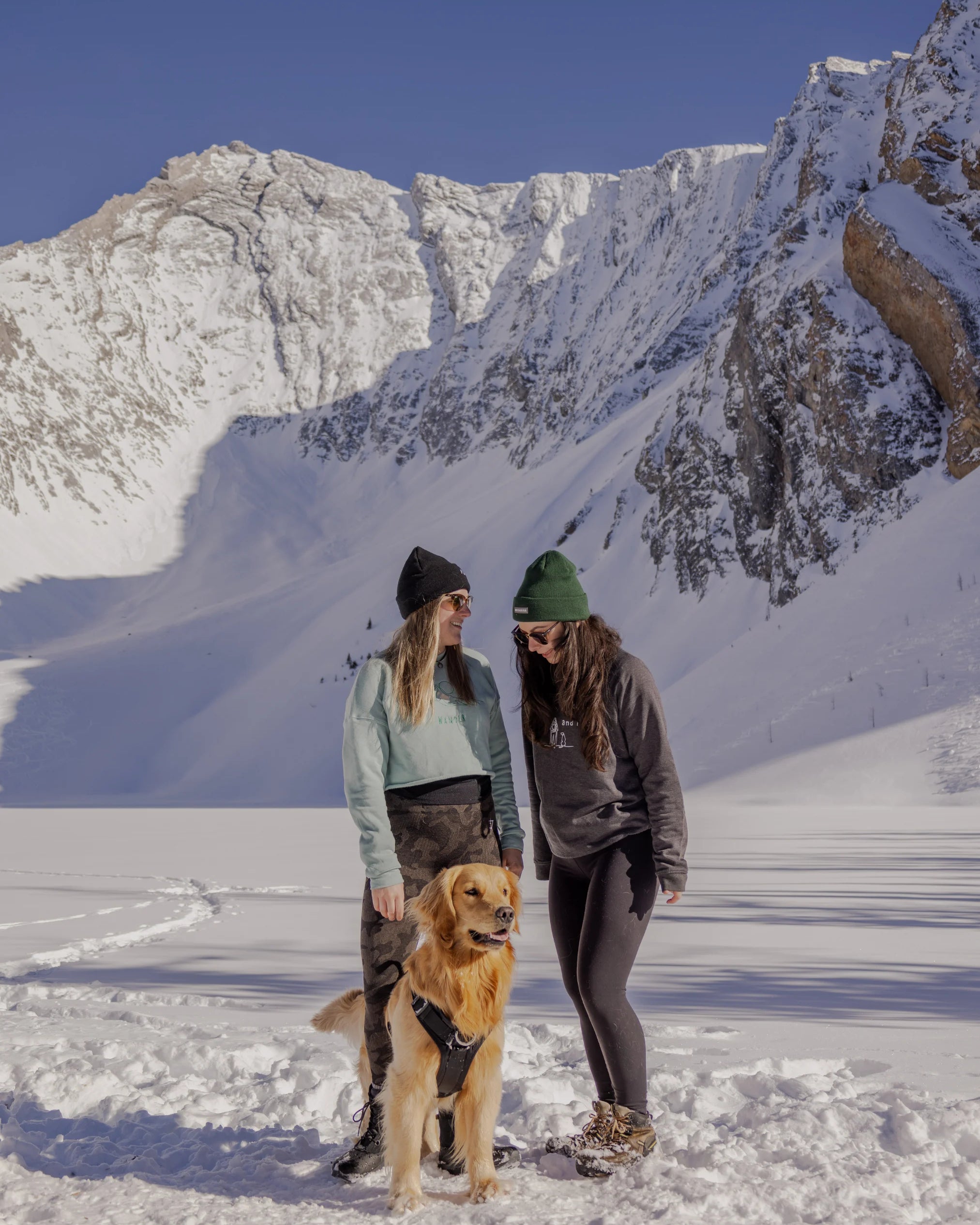 Two women wearing Nexarina gear and beanies, standing in the snow with a golden retriever in front of a dramatic alpine backdrop.