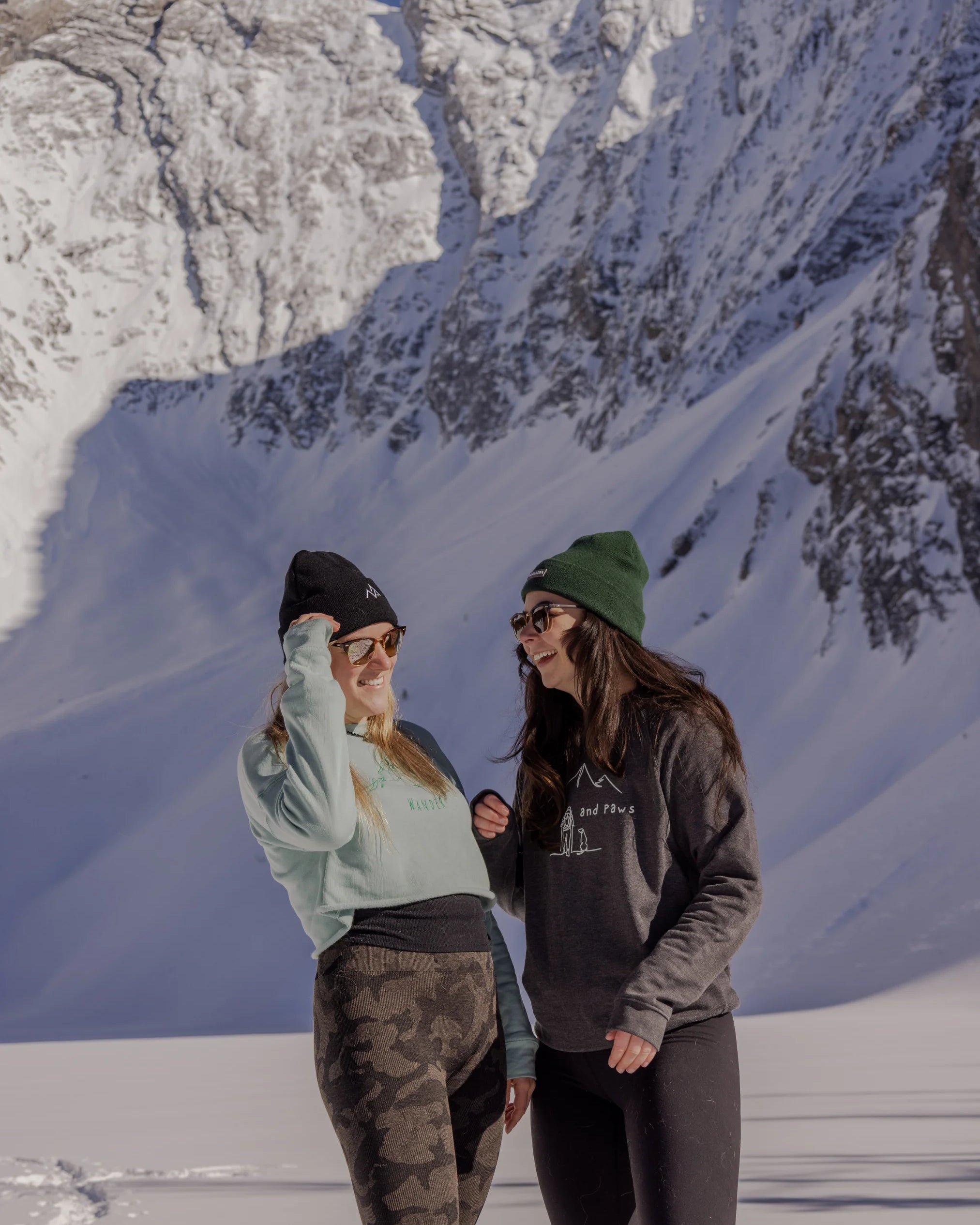 Two women in snow boots and Nexarina gear standing in a snowy valley, laughing with the mountains behind them.