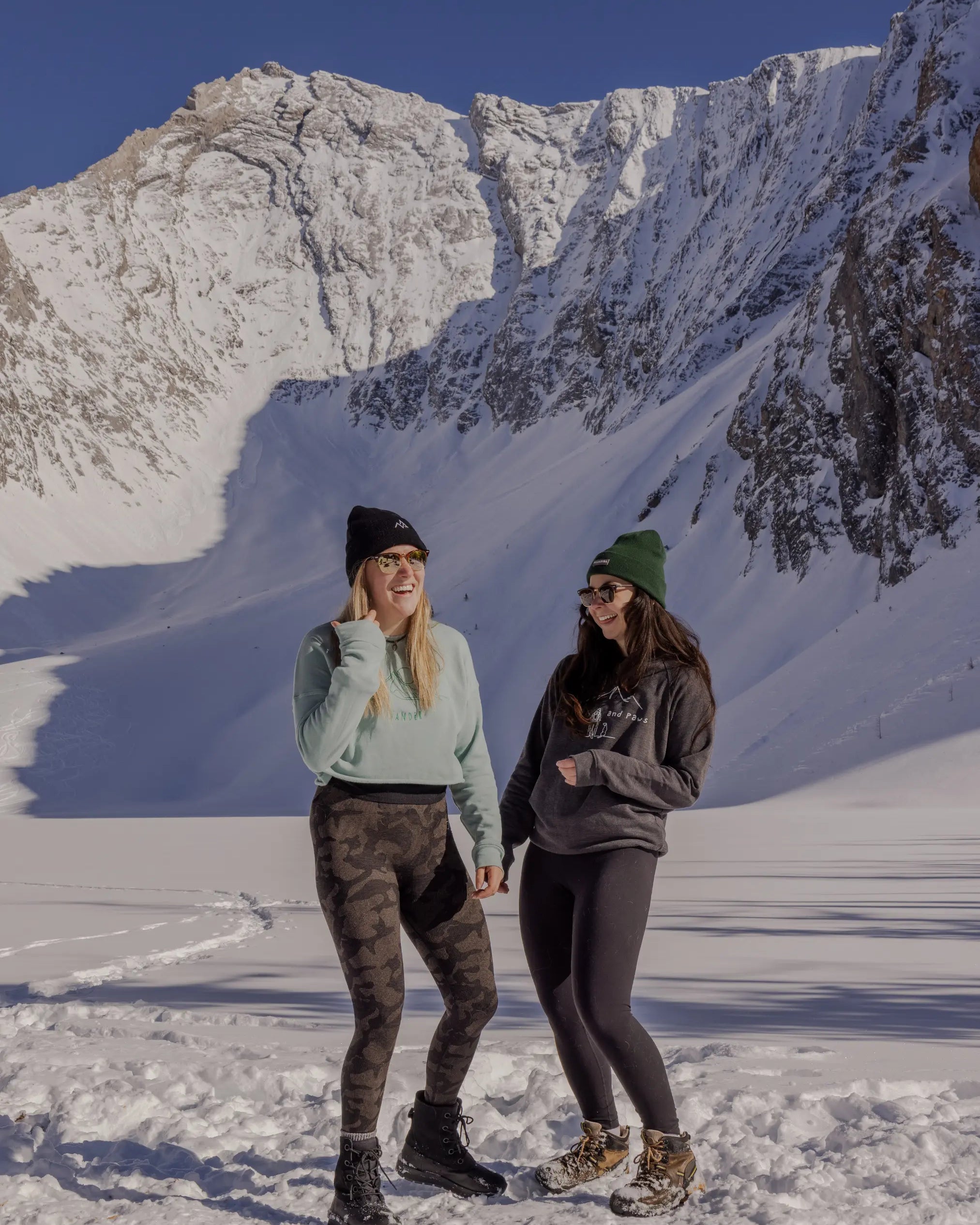 Two women in Nexarina winter apparel and beanies laughing in front of a steep snowy mountain face.