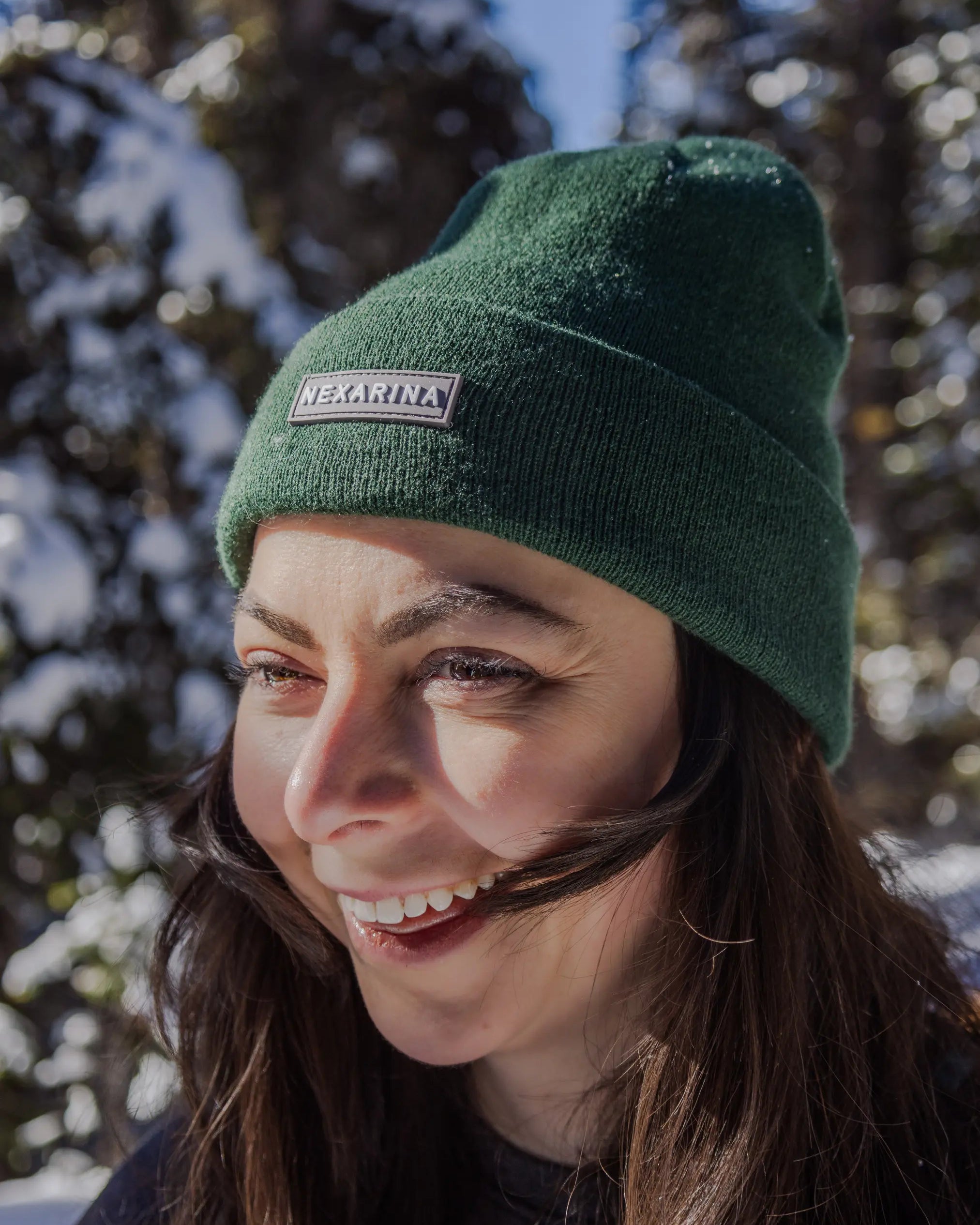 Close-up of a smiling woman in a Nexarina beanie, with trees and sunshine in the background. Her hair is catching the light.