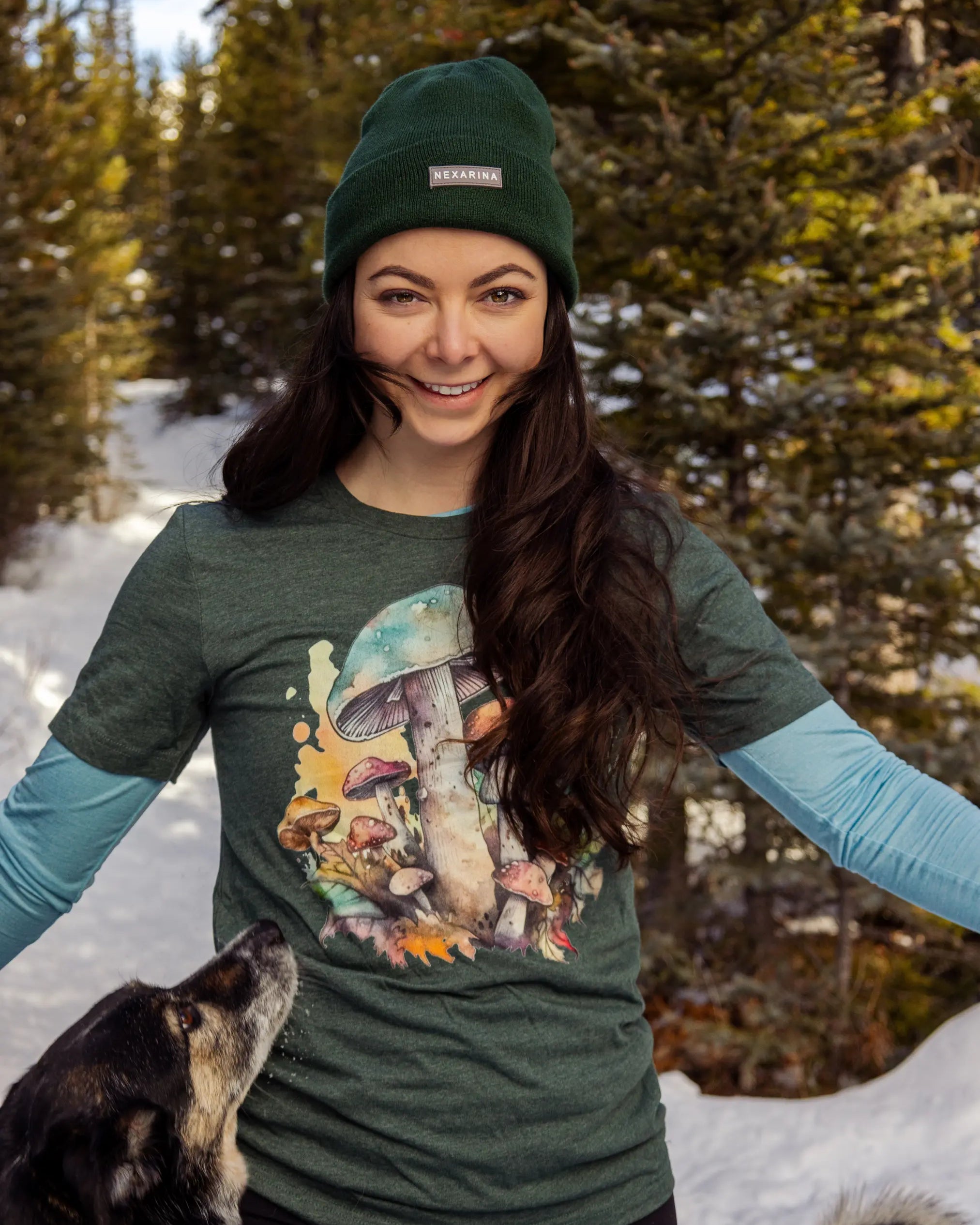 Woman wearing a forest green Nexarina beanie and a mushroom graphic tee, smiling on a snowy trail with a dog looking up at her.