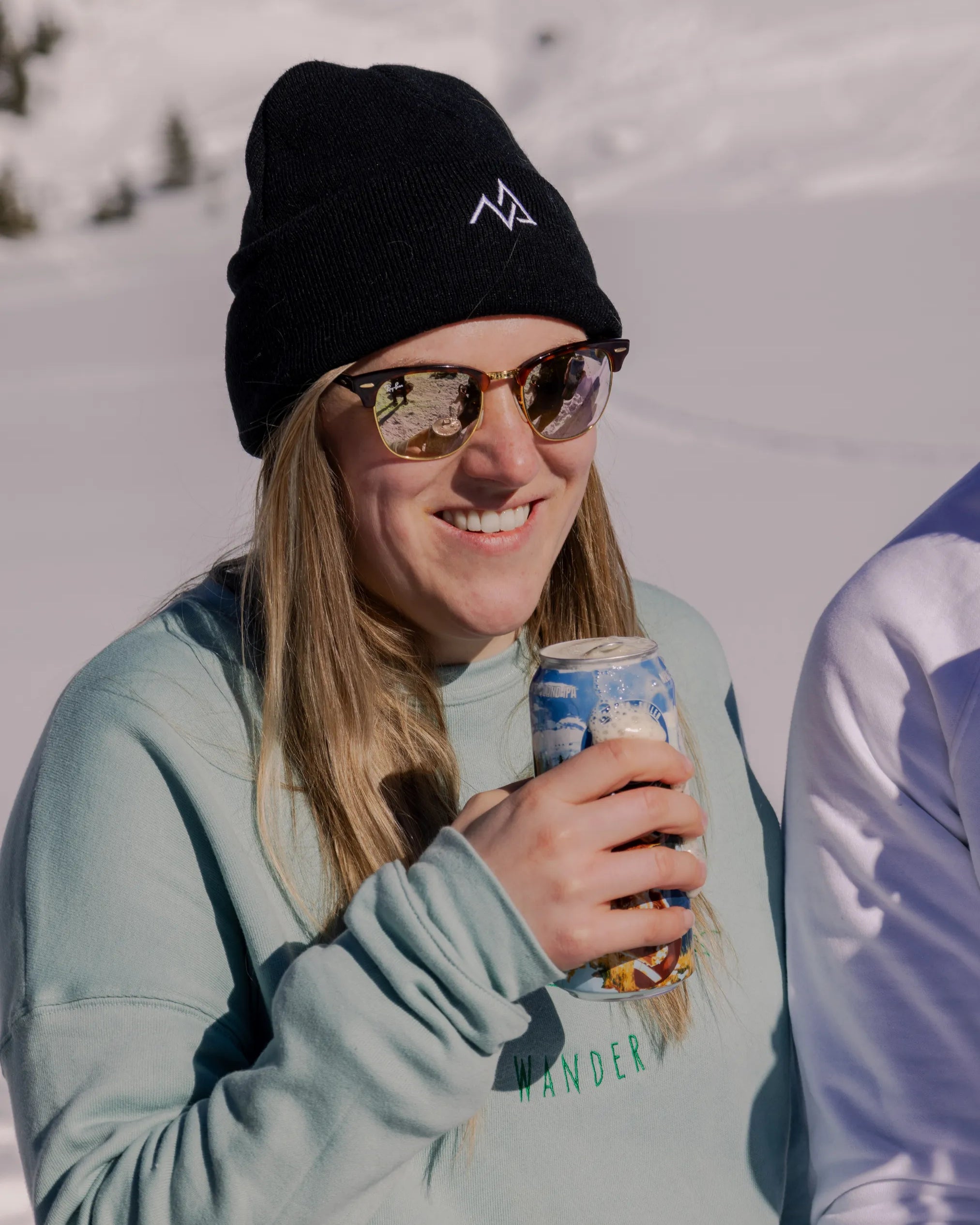 Woman wearing a black Nexarina beanie with embroidered logo, smiling in the sun with snowy mountains behind her.