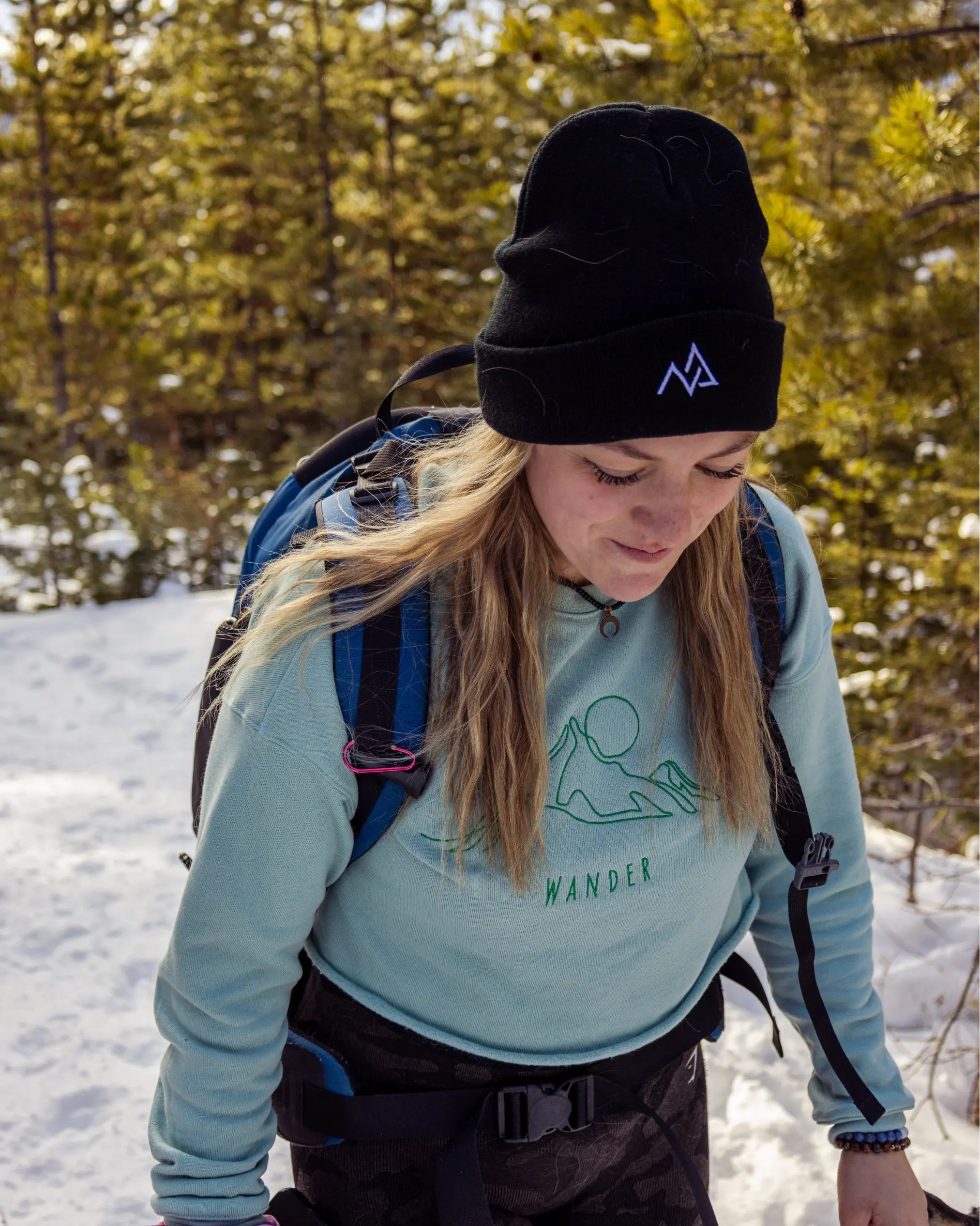 Close-up of a woman hiking in a black Nexarina beanie and mint green crewneck, trekking through a snowy forest.