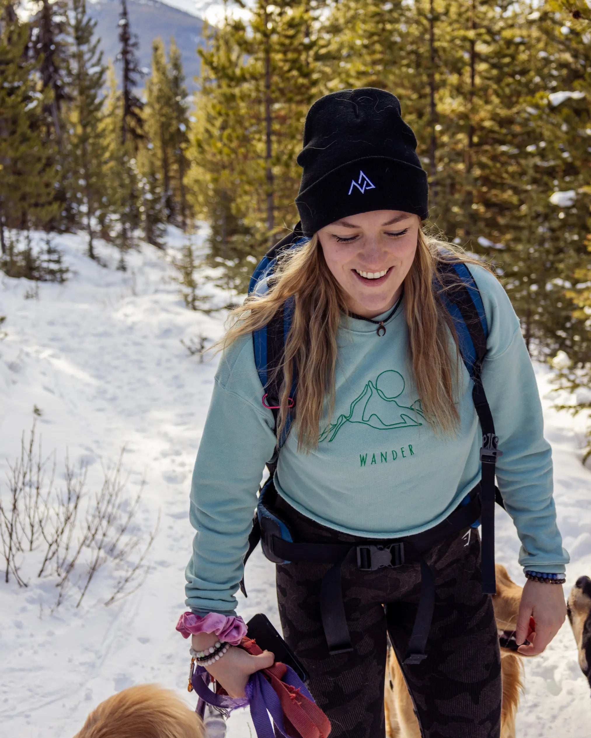 Woman wearing a black Nexarina beanie, smiling while hiking snowy trails with dogs around her.