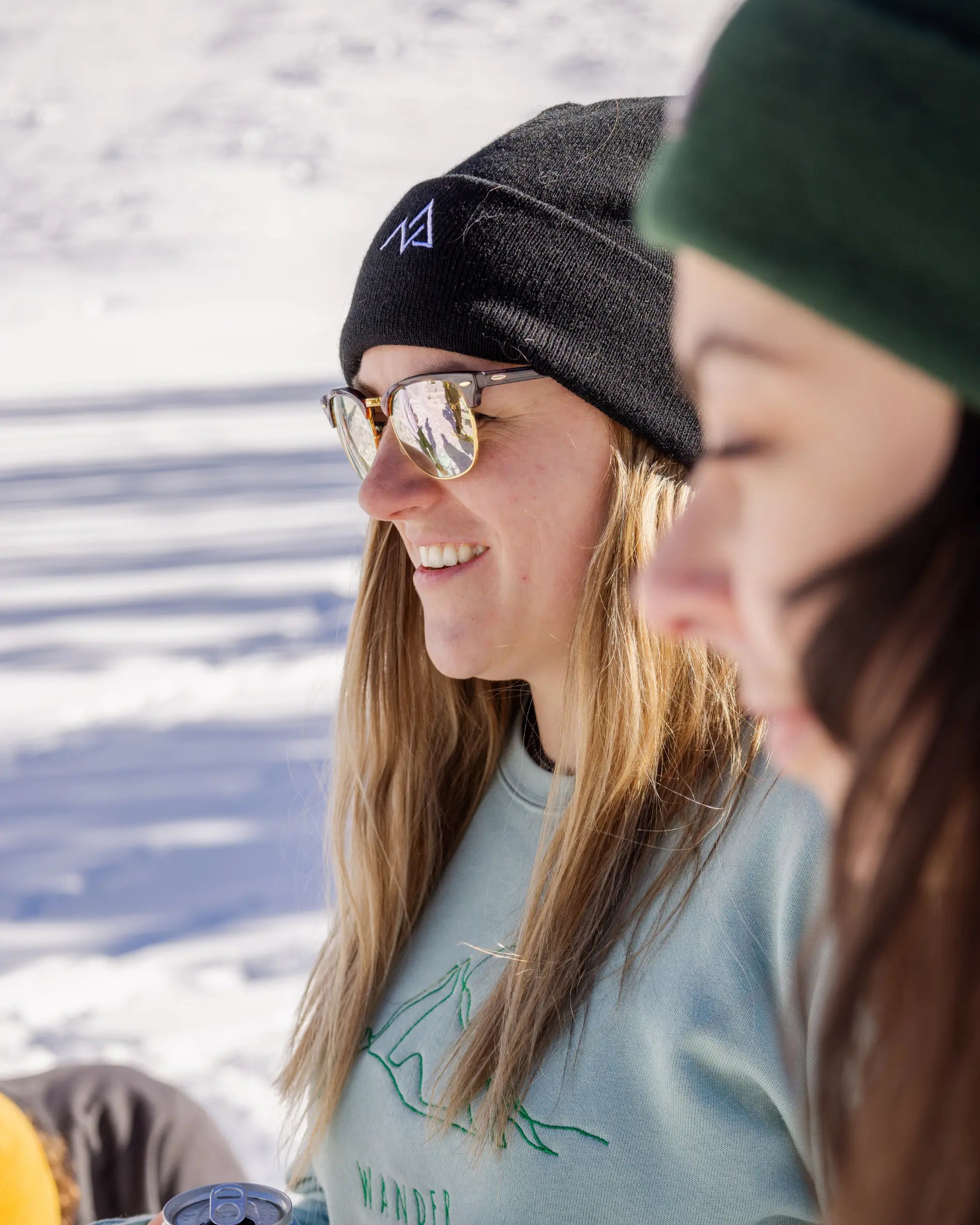 Side profile of a woman in a black Nexarina beanie and reflective sunglasses, holding a drink and smiling outdoors.
