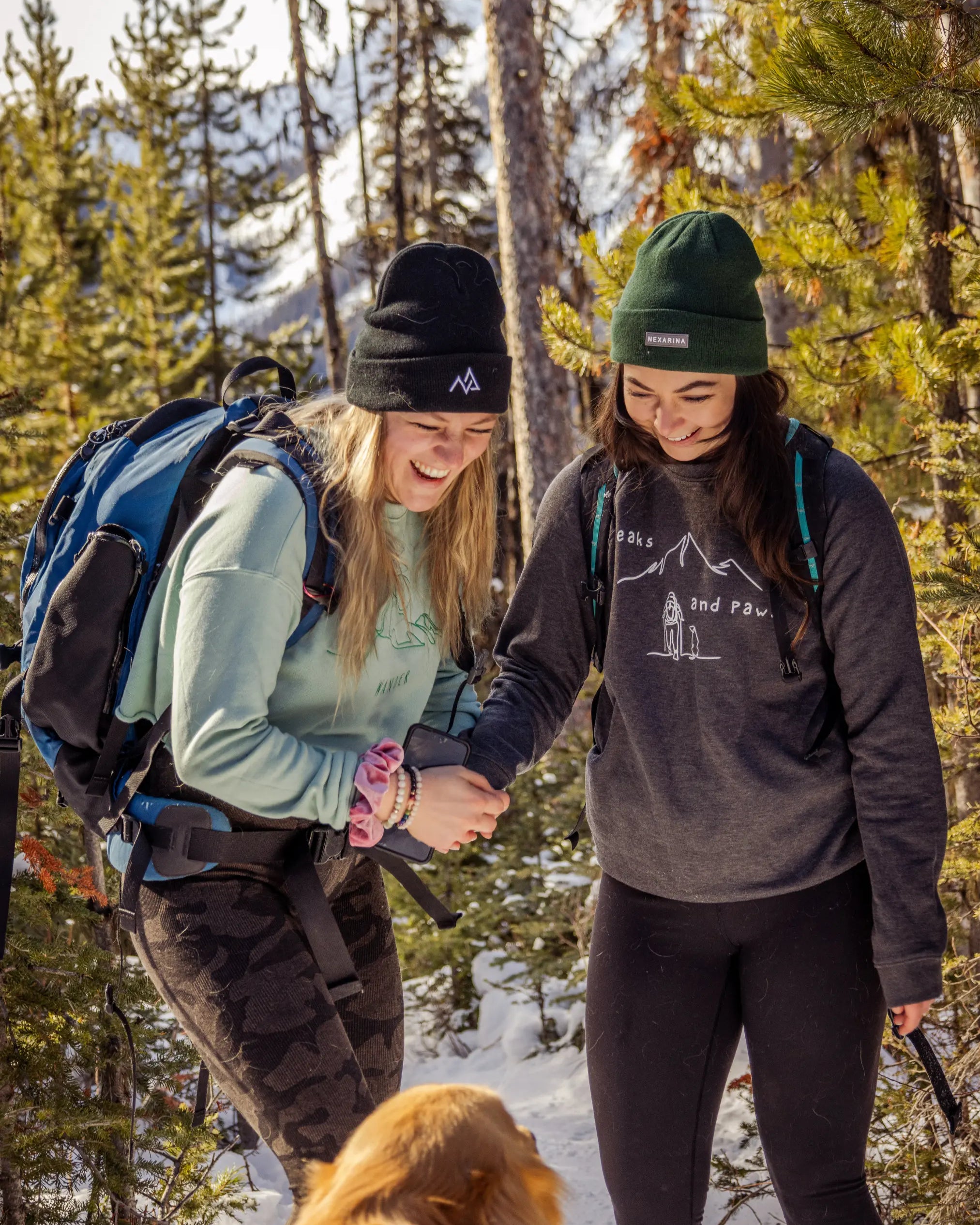 Two women laughing in the forest, one wearing a black Nexarina beanie and the other in green, holding hands and surrounded by snow and pine trees.