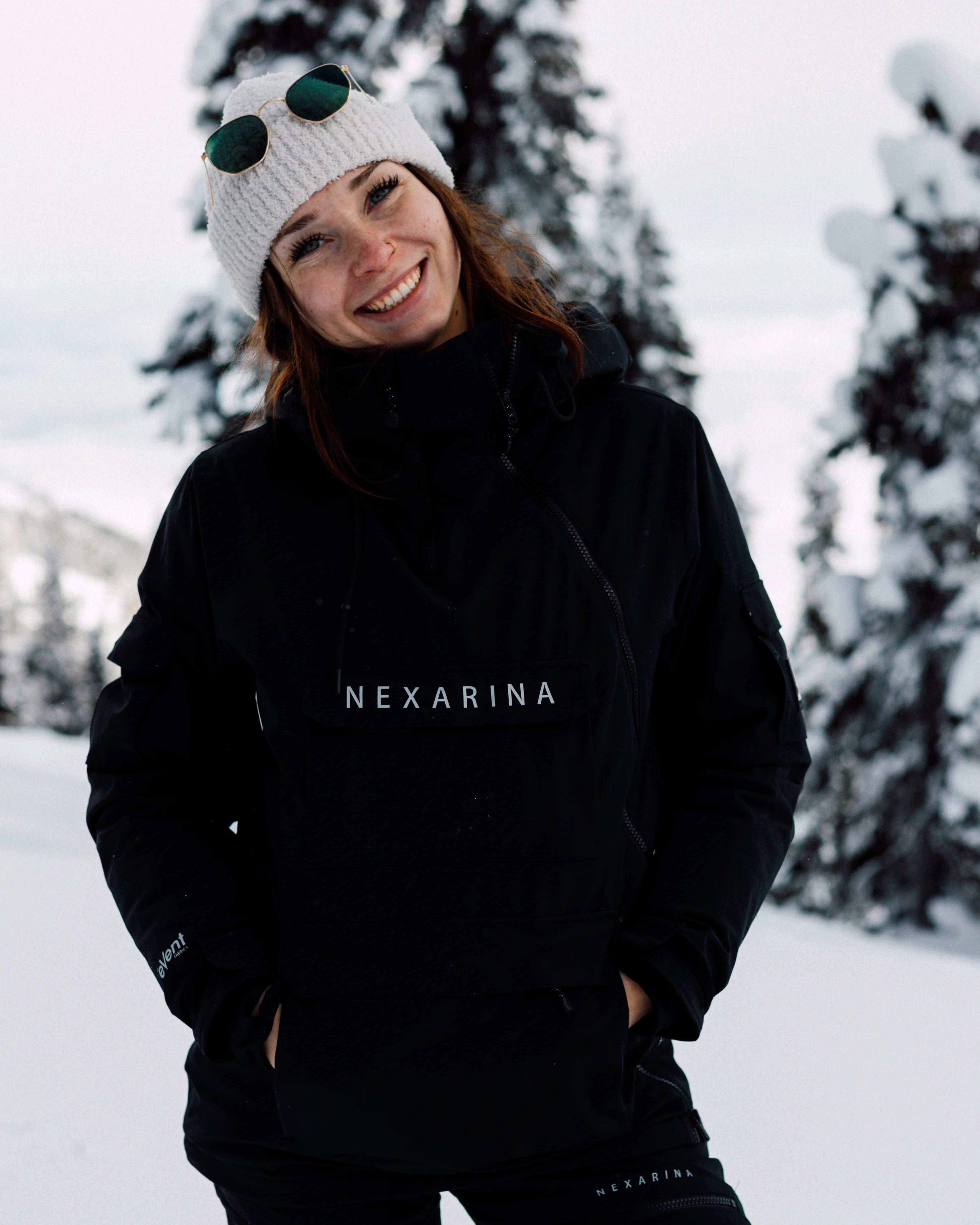 Woman smiling in snowy forest wearing Nexarina Indy Snow Jacket and branded beanie.