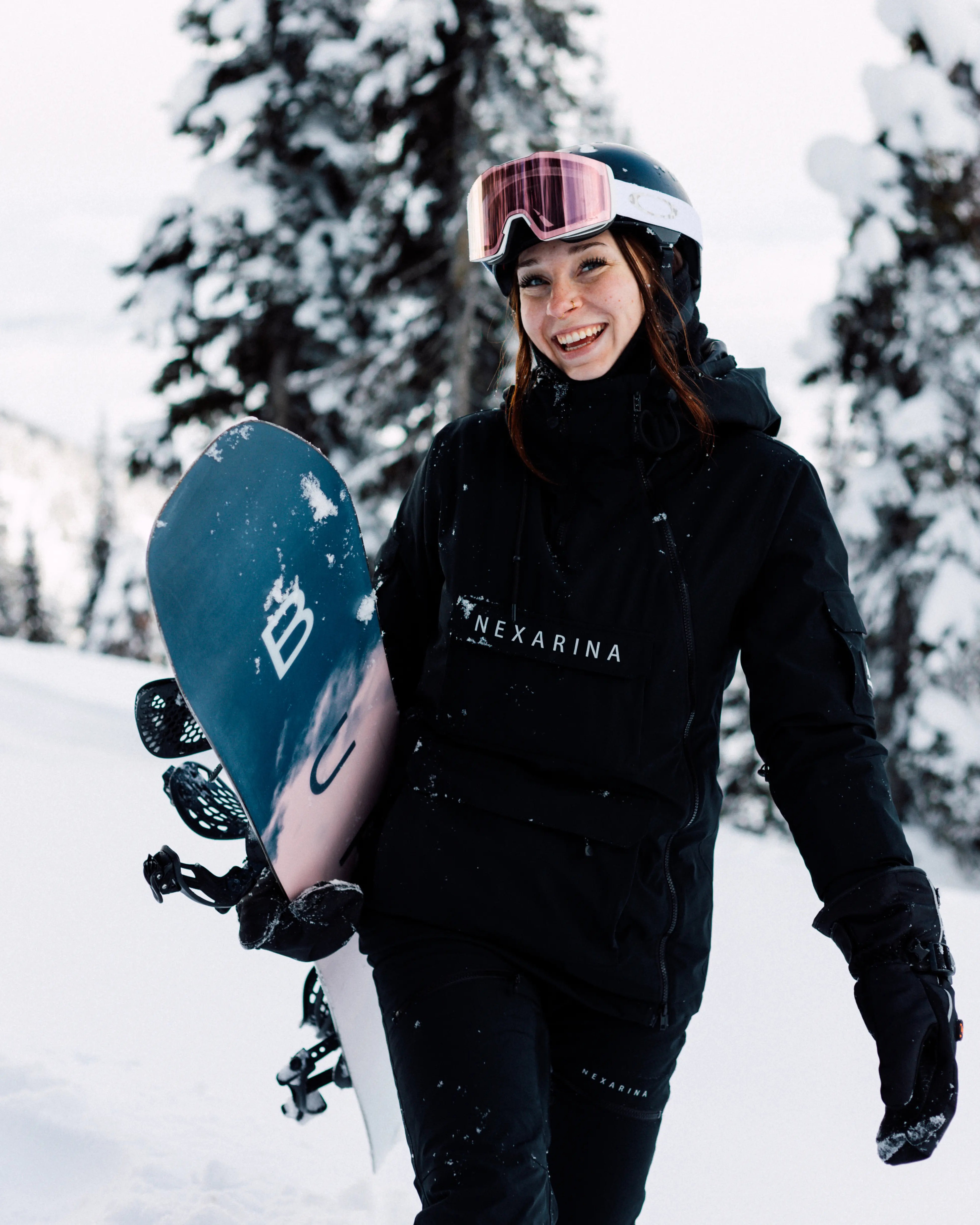 Woman smiling while holding snowboard in snowy setting, wearing Nexarina Indy Snow Jacket and pants