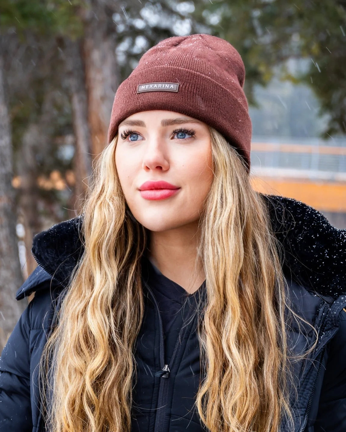 Portrait of a young woman wearing Nexarina Coco beanie, snow falling gently around her.