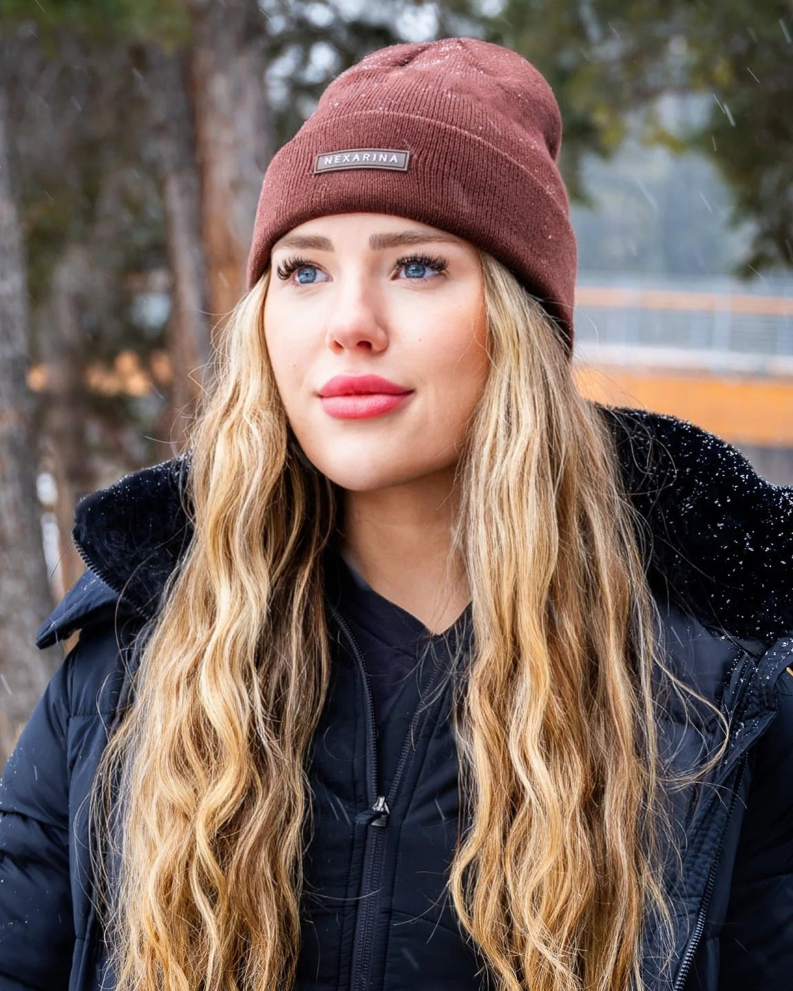 Portrait of a young woman wearing Nexarina Coco beanie, snow falling gently around her.