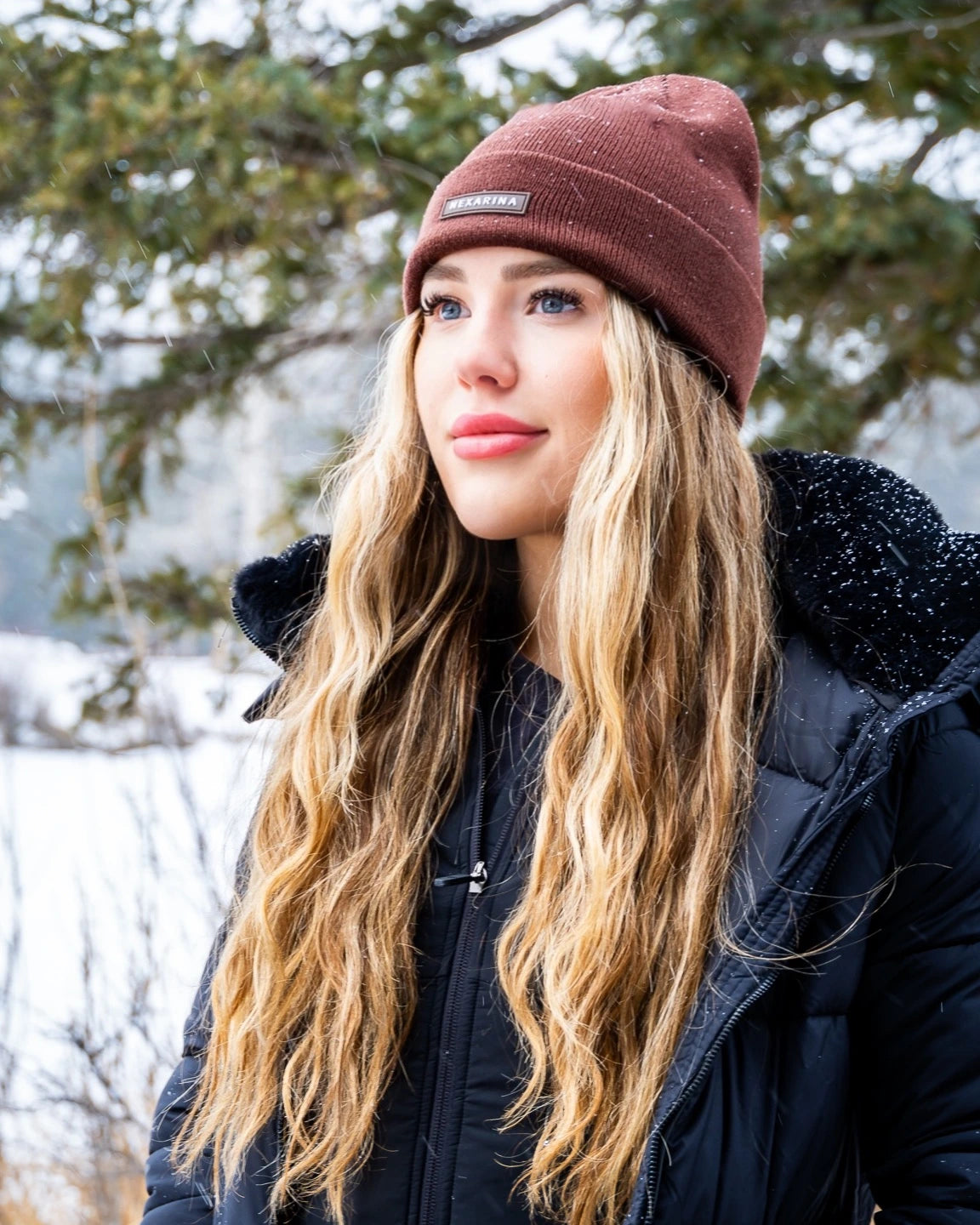 Woman in black jacket and Nexarina Coco brown beanie standing in snowy forest.