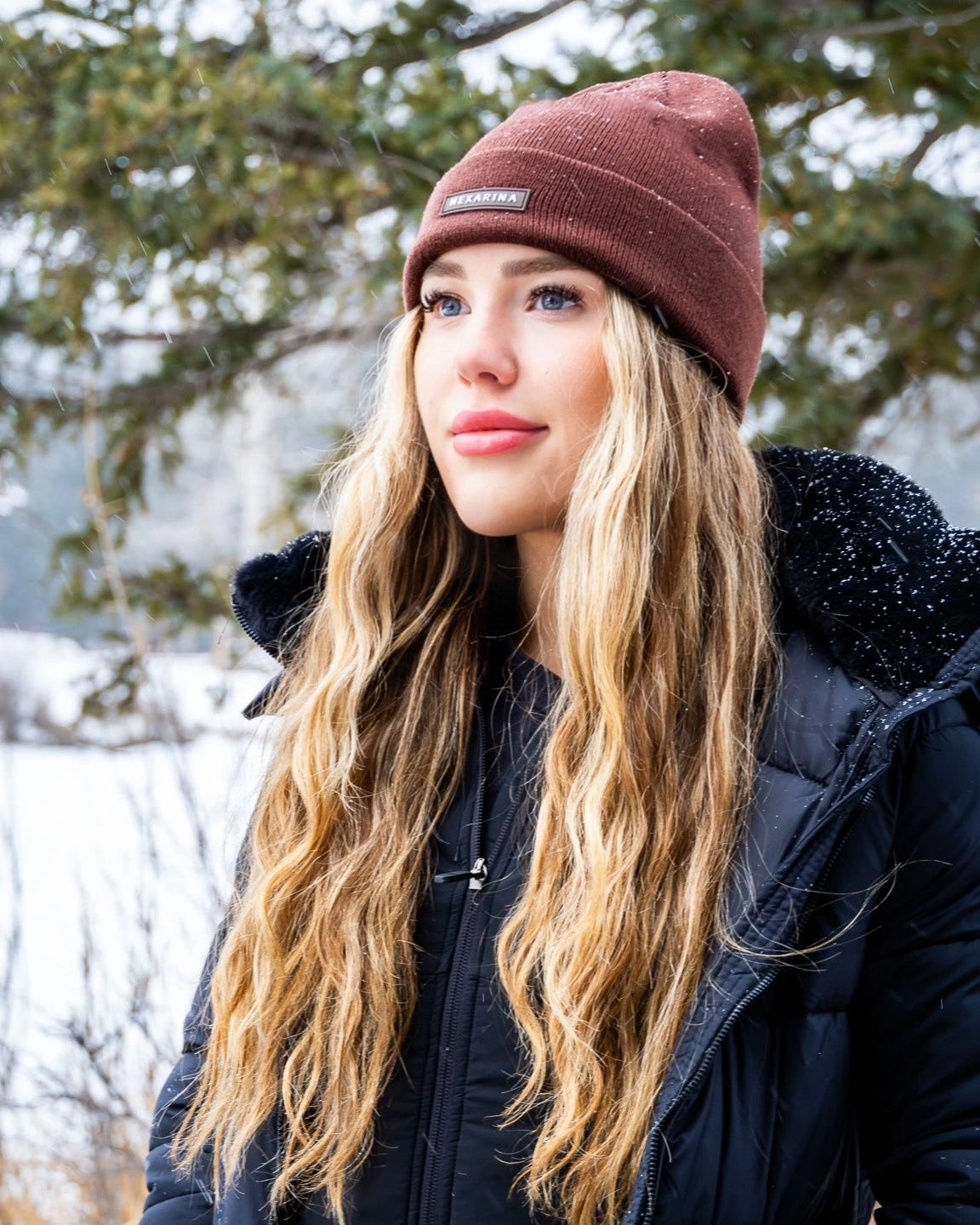 Woman in black jacket and Nexarina Coco brown beanie standing in snowy forest.
