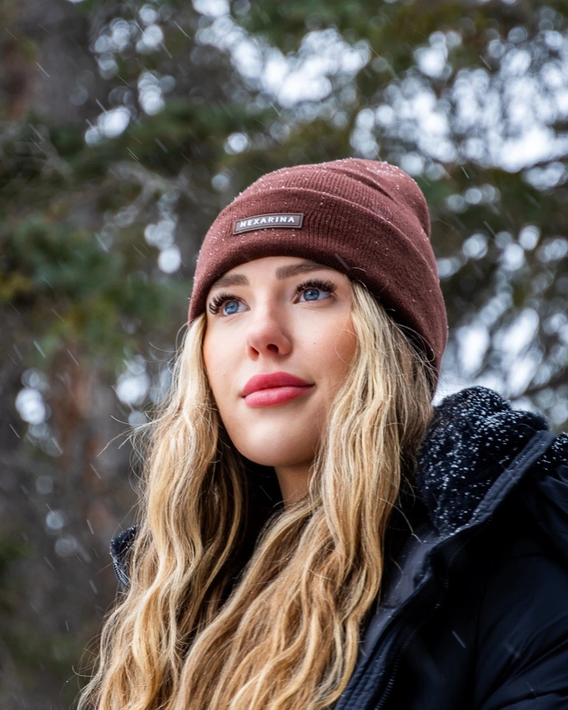 Close-up of a young woman in a Nexarina Coco brown beanie, snow gently falling around her.