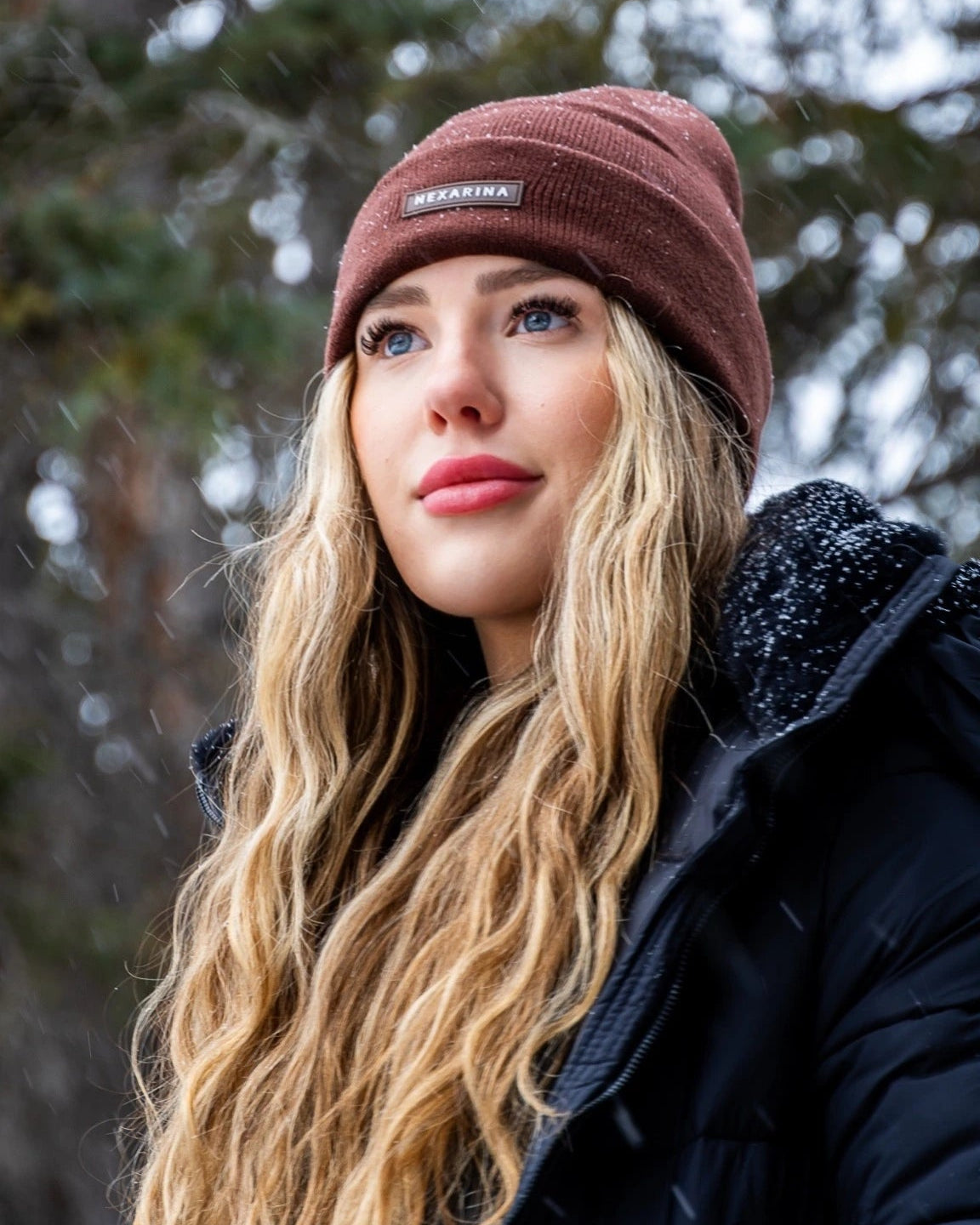 Close-up of a young woman in a Nexarina Coco brown beanie, snow gently falling around her.