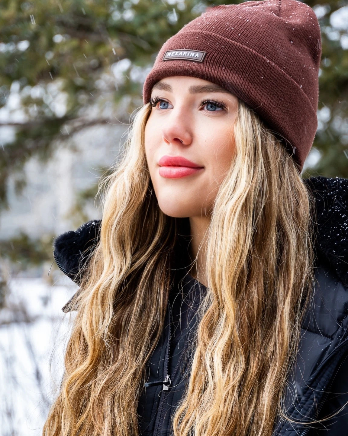 Blonde woman wearing Nexarina Coco beanie in brown, looking thoughtfully in a snowy setting.
