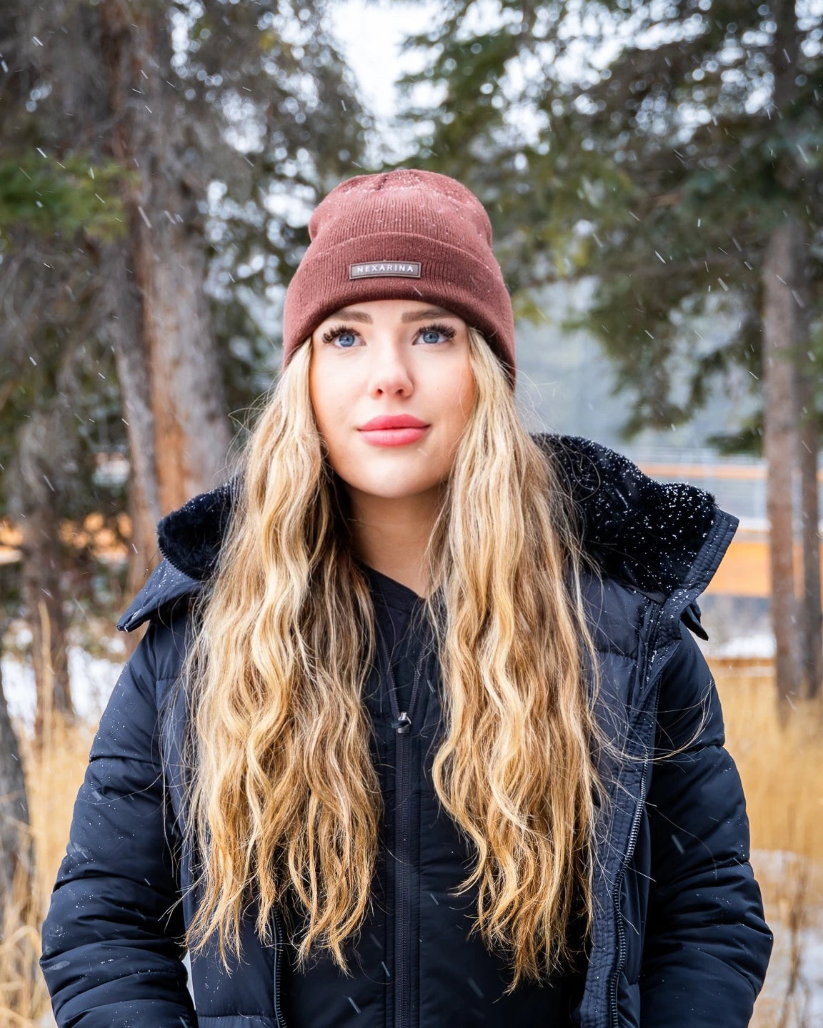 Blonde woman in Nexarina Coco beanie with a thoughtful gaze, snowy trees in the background.