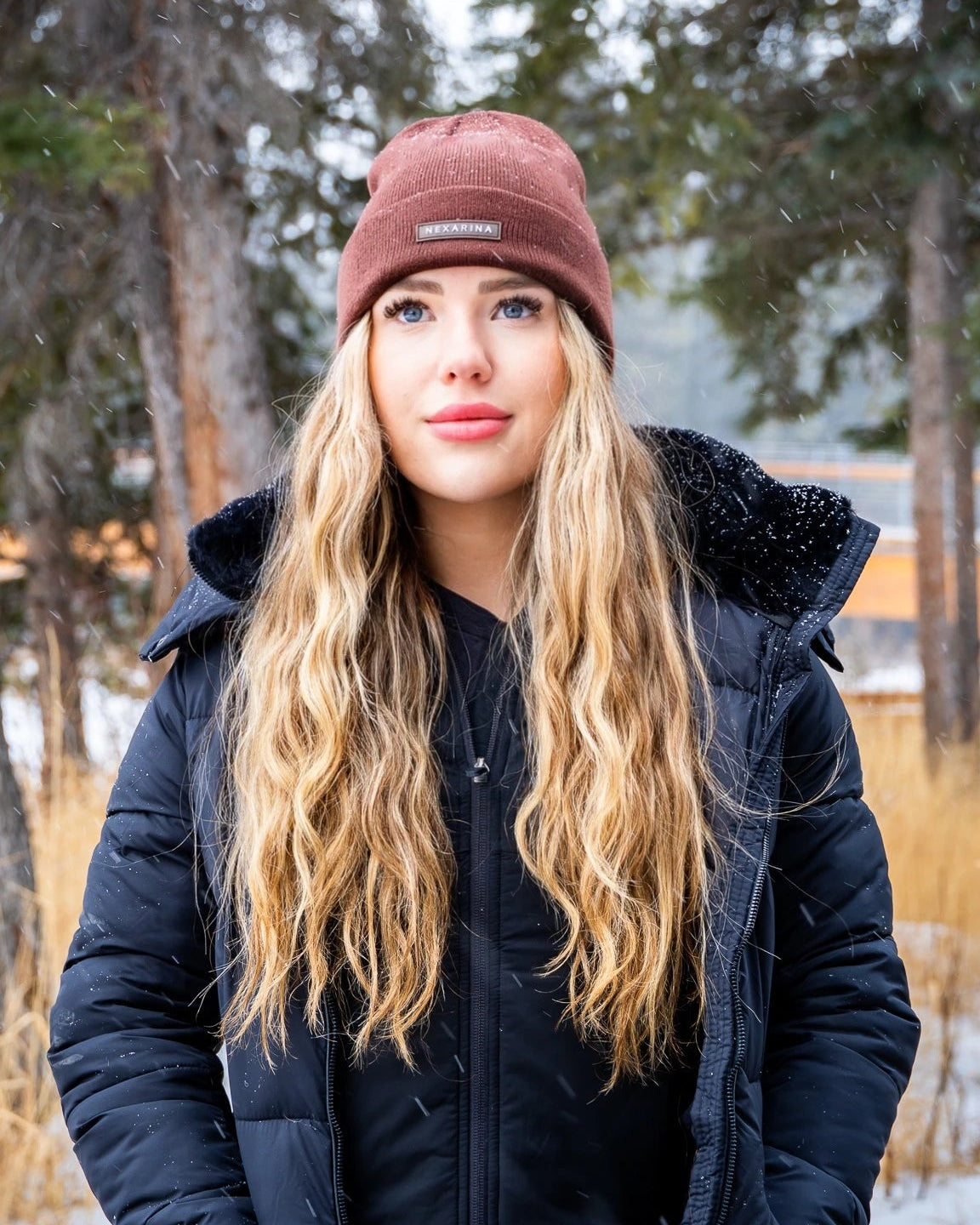 Blonde woman in Nexarina Coco beanie with a thoughtful gaze, snowy trees in the background.