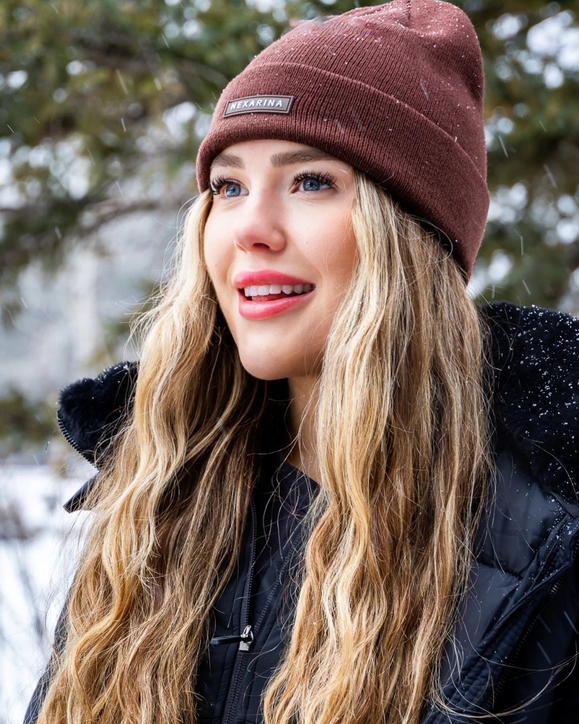 Smiling woman outdoors wearing Nexarina Coco brown beanie, with snow-covered trees in the background.