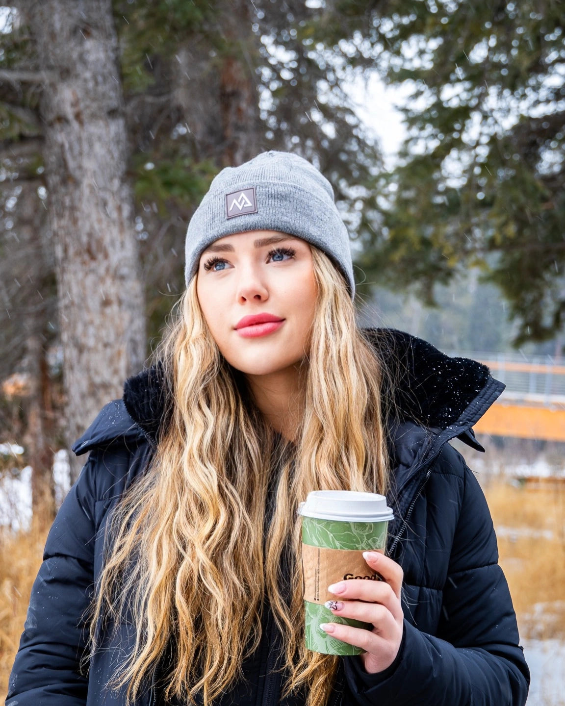 Winter portrait of a woman in a Nexarina grey beanie, enjoying a hot drink in a snowy woodland.