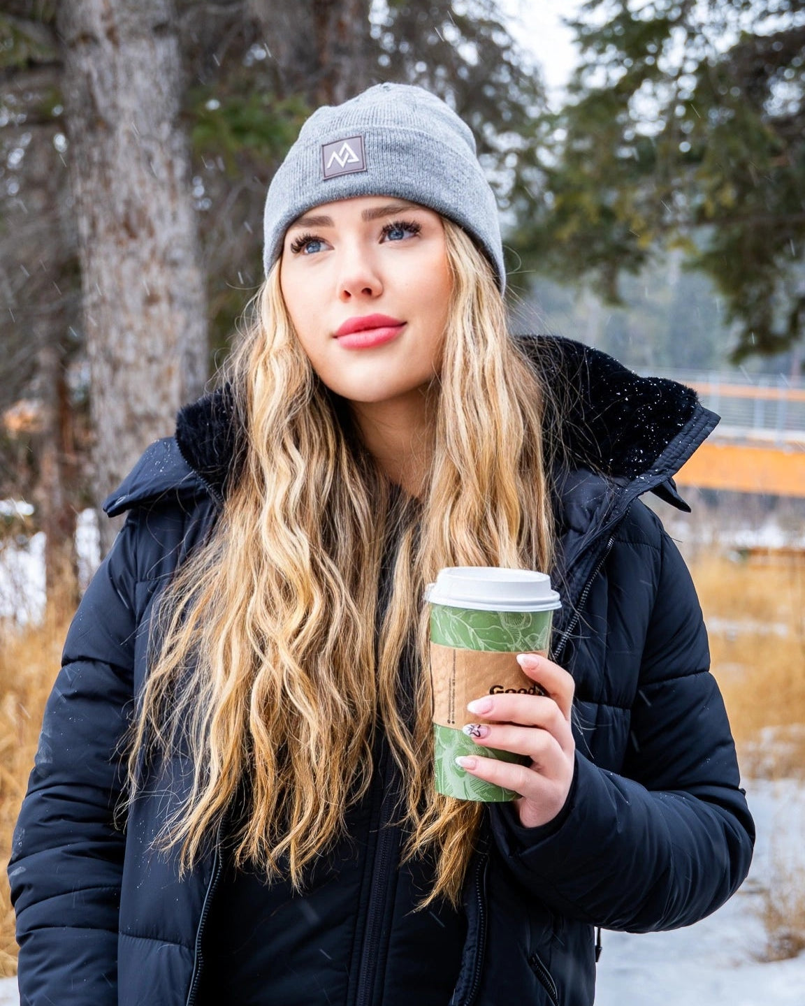 Winter portrait of a woman in a Nexarina grey beanie, enjoying a hot drink in a snowy woodland.