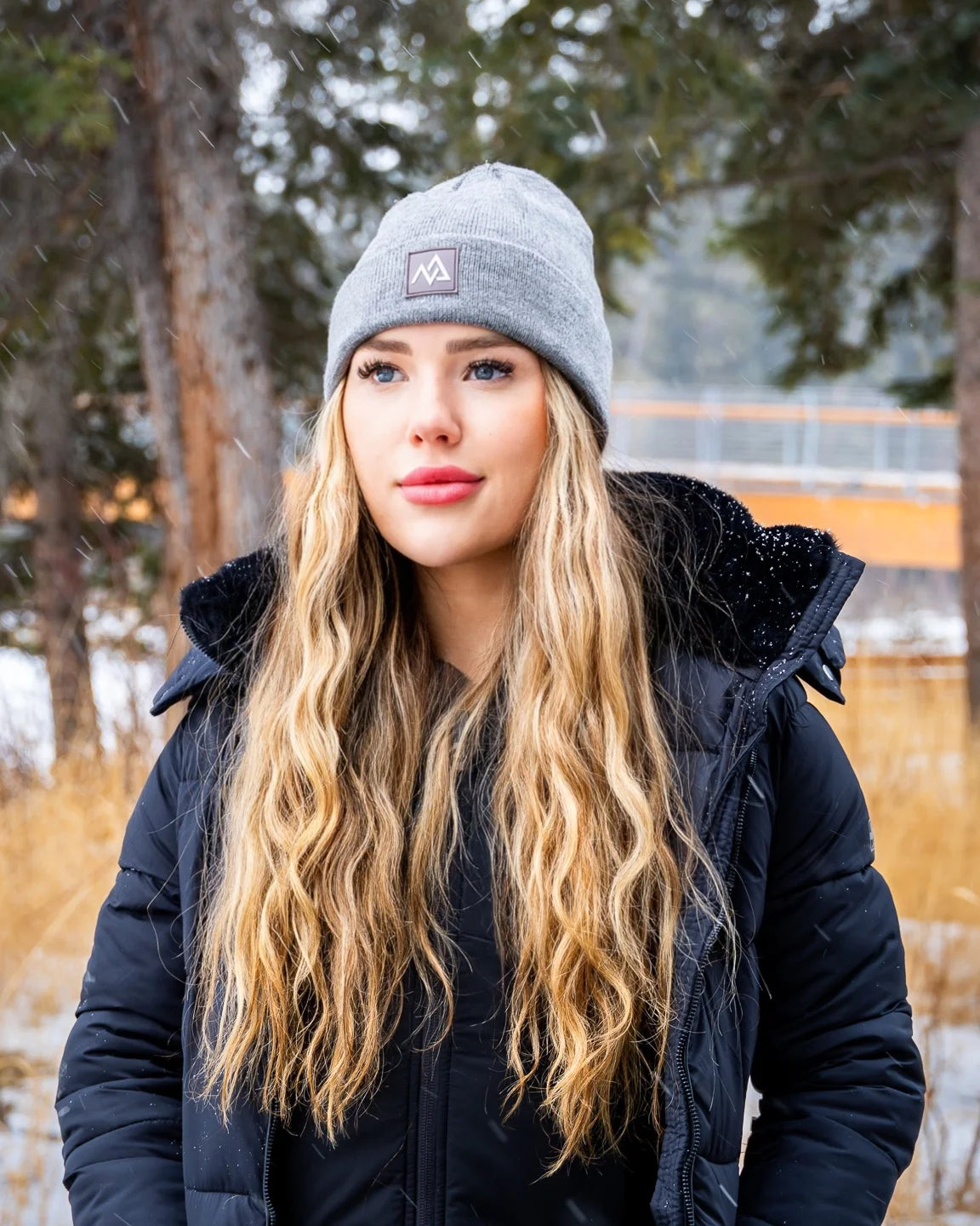 Woman in a grey Nexarina beanie and black coat, smiling gently in a snowy forest setting.