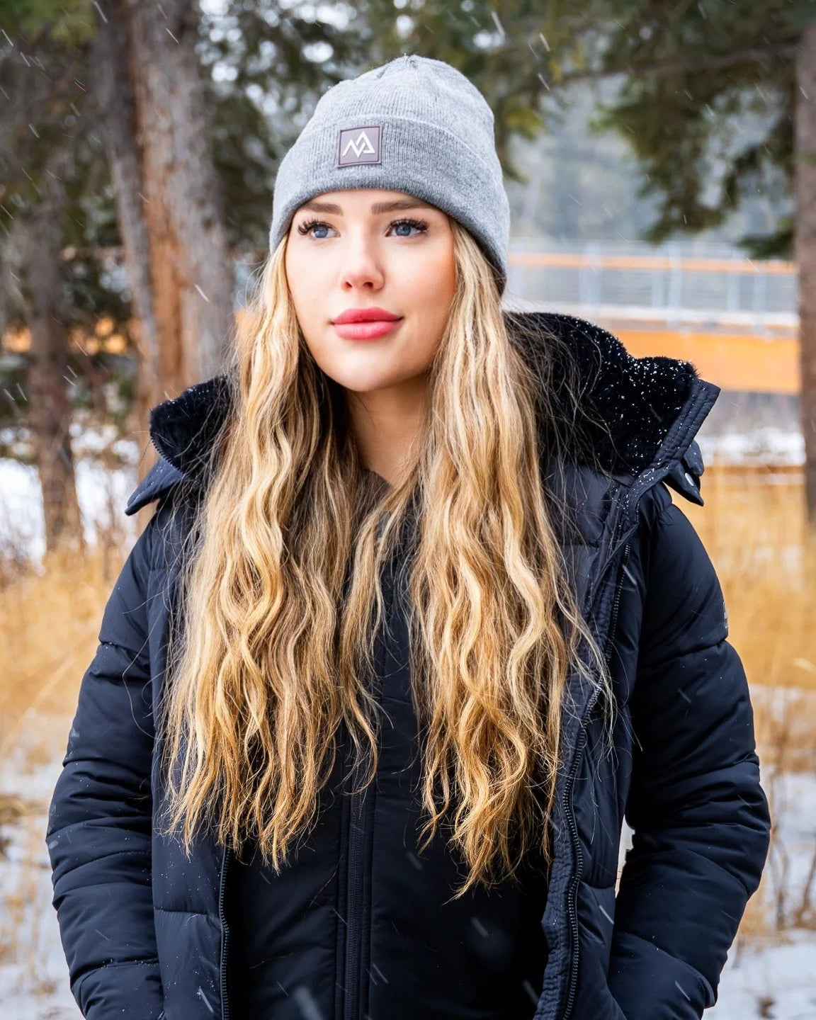 Woman in a grey Nexarina beanie and black coat, smiling gently in a snowy forest setting.