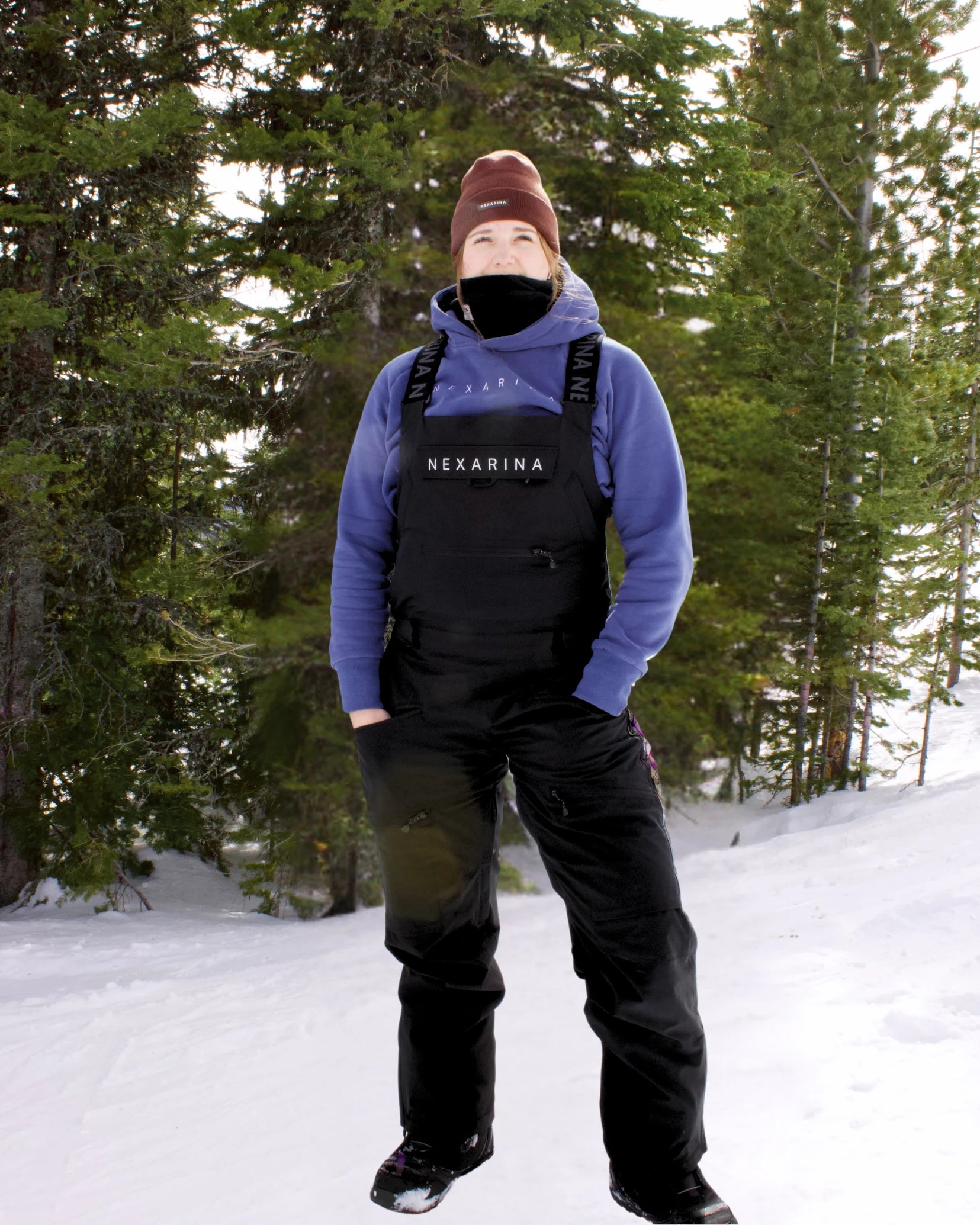 Woman standing in the snow wearing the Nexarina Everette Bib in black, paired with a Nexarina hoodie and the Coco Beanie. The scene shows a forest backdrop with snow-covered ground, highlighting the functional fit and design of Nexarina women’s outerwear.