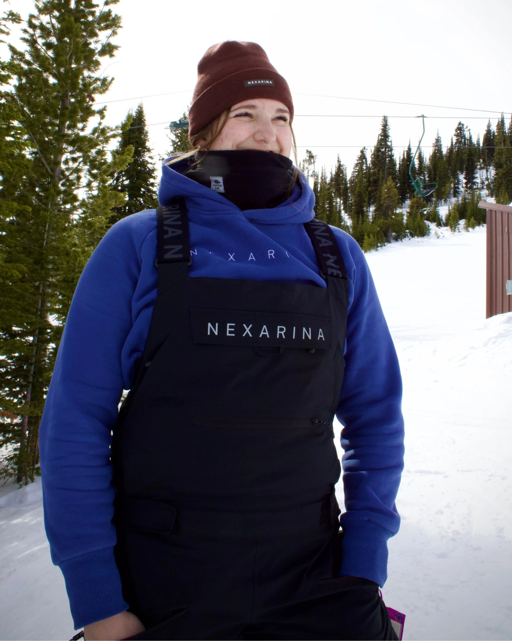 Woman standing in the snow wearing Nexarina Everette Bib black snowboard overall.