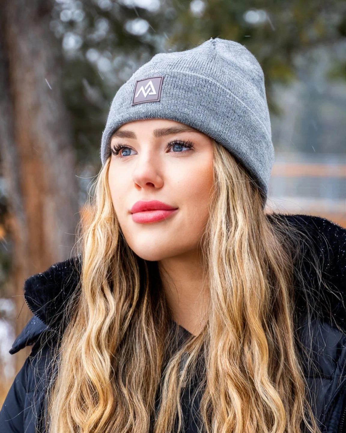 Close-up of a woman wearing a grey Nexarina Emery Beanie, set against a winter forest backdrop.