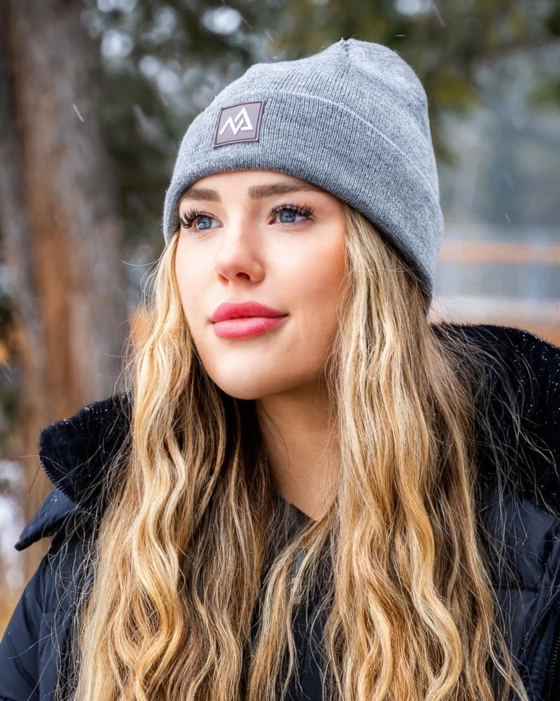 Close-up of a woman wearing a grey Nexarina Emery Beanie, set against a winter forest backdrop.