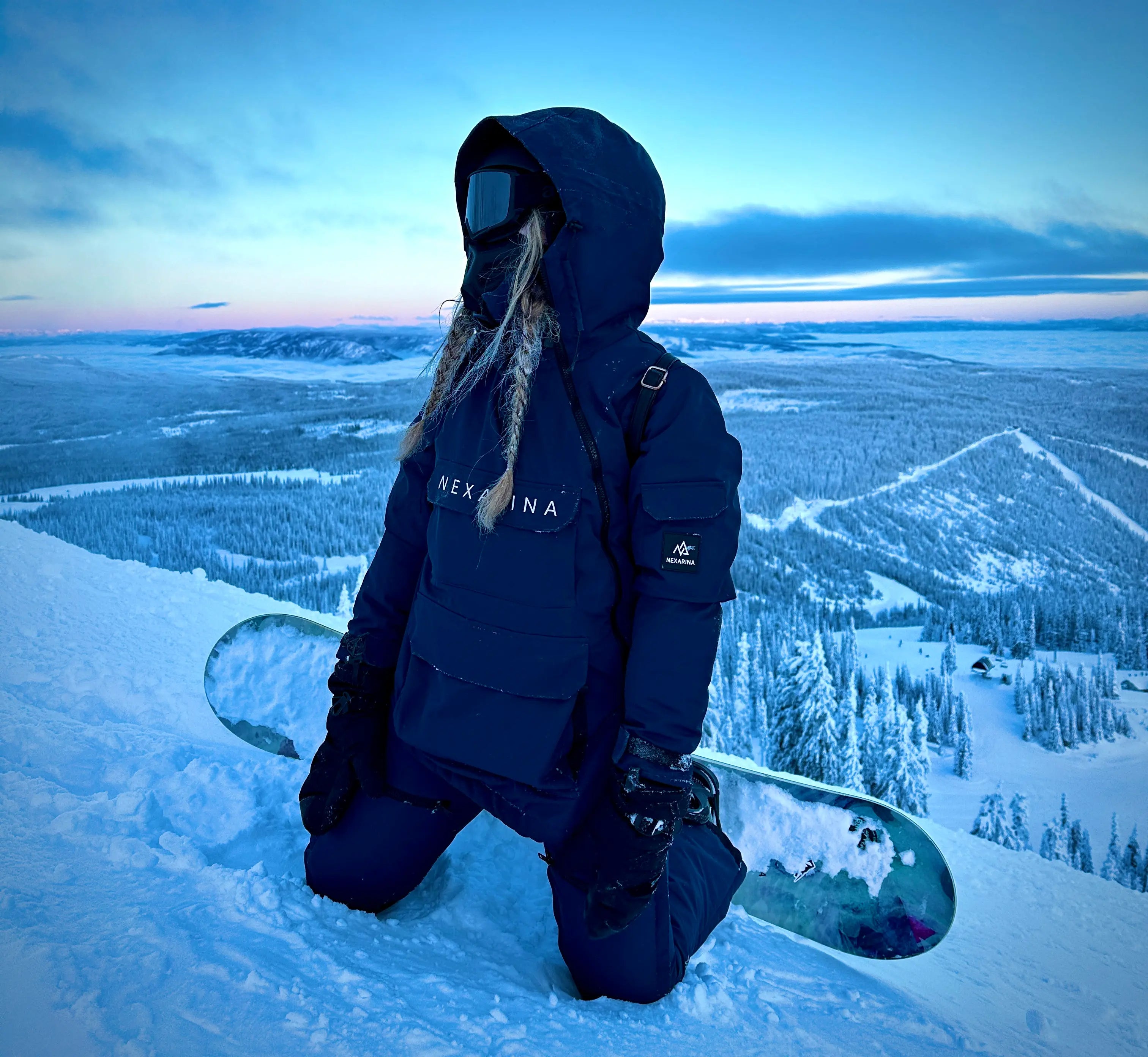 Woman kneeling on a snowy mountain in full Nexarina snow gear, wearing a black jacket and bib with goggles and a snowboard beside her. Captured at sunset overlooking a vast winter landscape, highlighting the performance and design of women’s technical outerwear.
