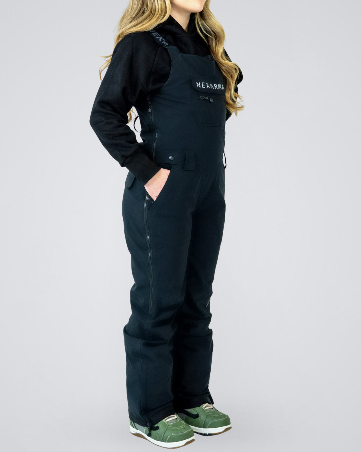 Woman wearing Nexarina Everette women’s black bib snow pants, side profile showing tailored fit and full-length leg coverage.