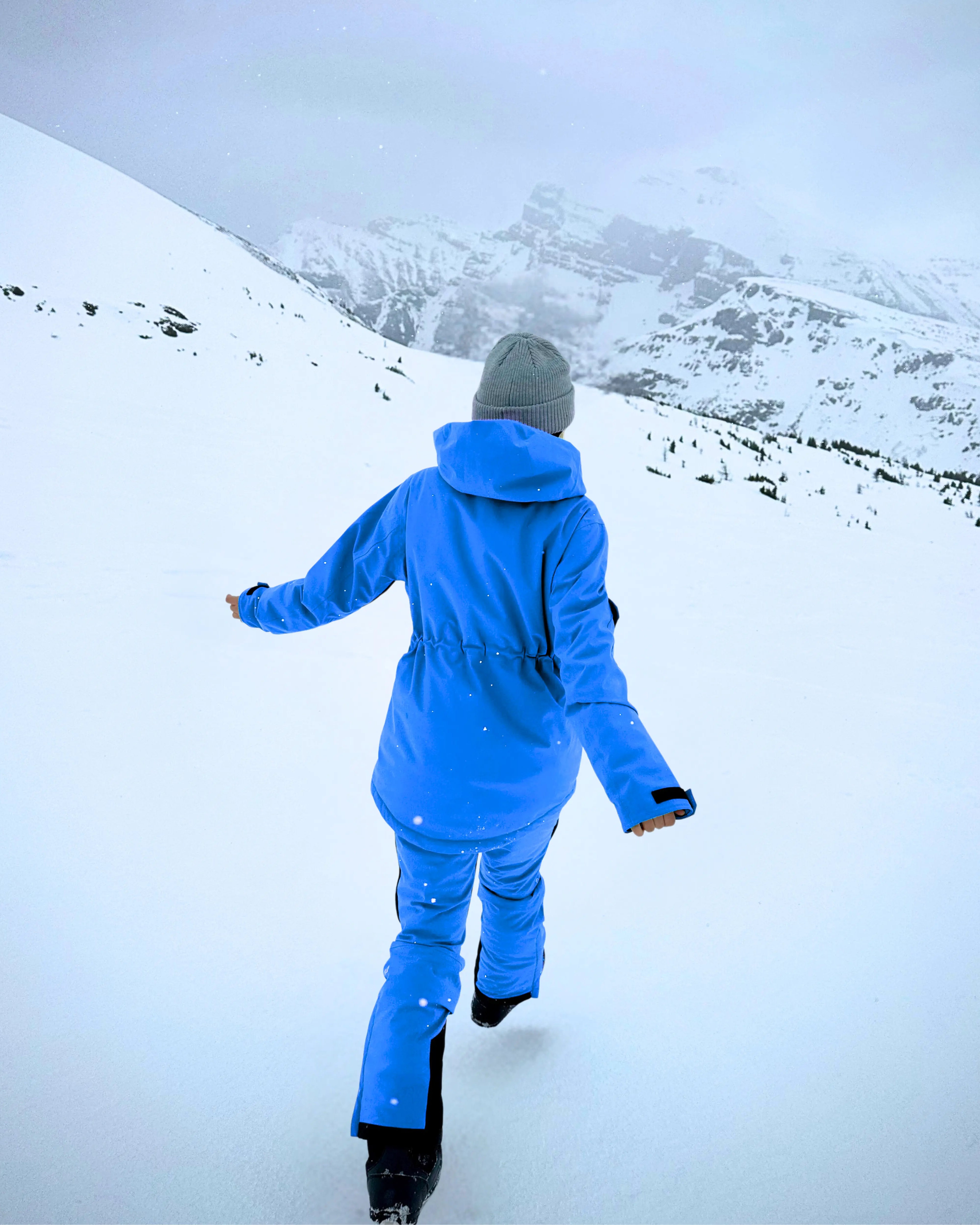A model walks through the snow in blue Avani snow pants, featuring an adjustable waist, insulation, and flexible design.