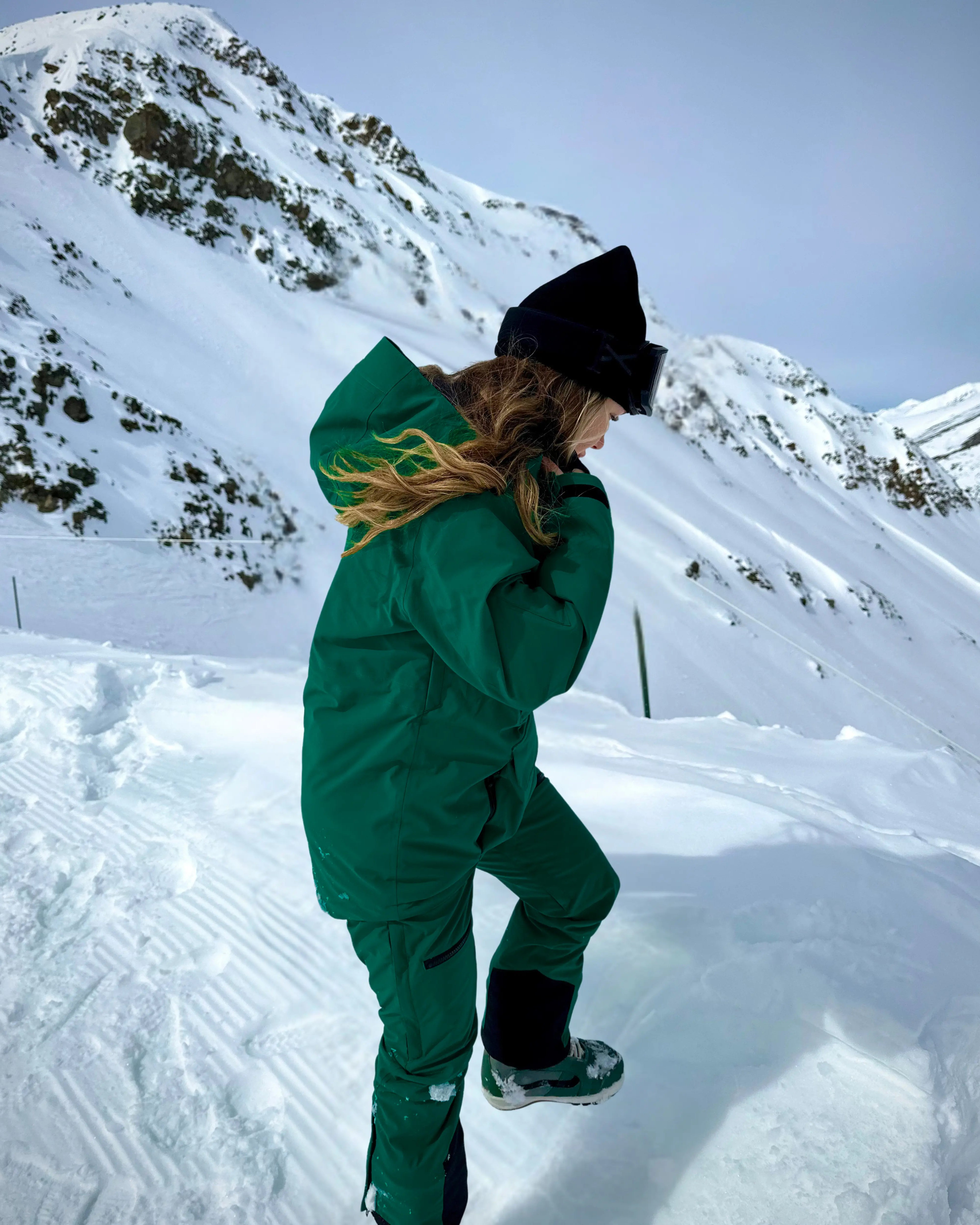 Woman wearing Nexarina Avani snowpants in emerald green standing in deep snow with mountain backdrop.