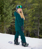 Woman wearing Nexarina women’s bib snow pants in emerald green, side view on a snowboard in alpine terrain.