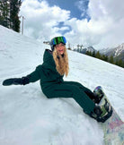 Woman wearing Nexarina emerald green bib snow pants, seated on snow with snowboard in a mountain setting.
