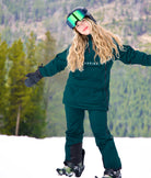 Front view of Nexarina women’s snow jacket and bib pants in emerald green on a snowy mountainside.