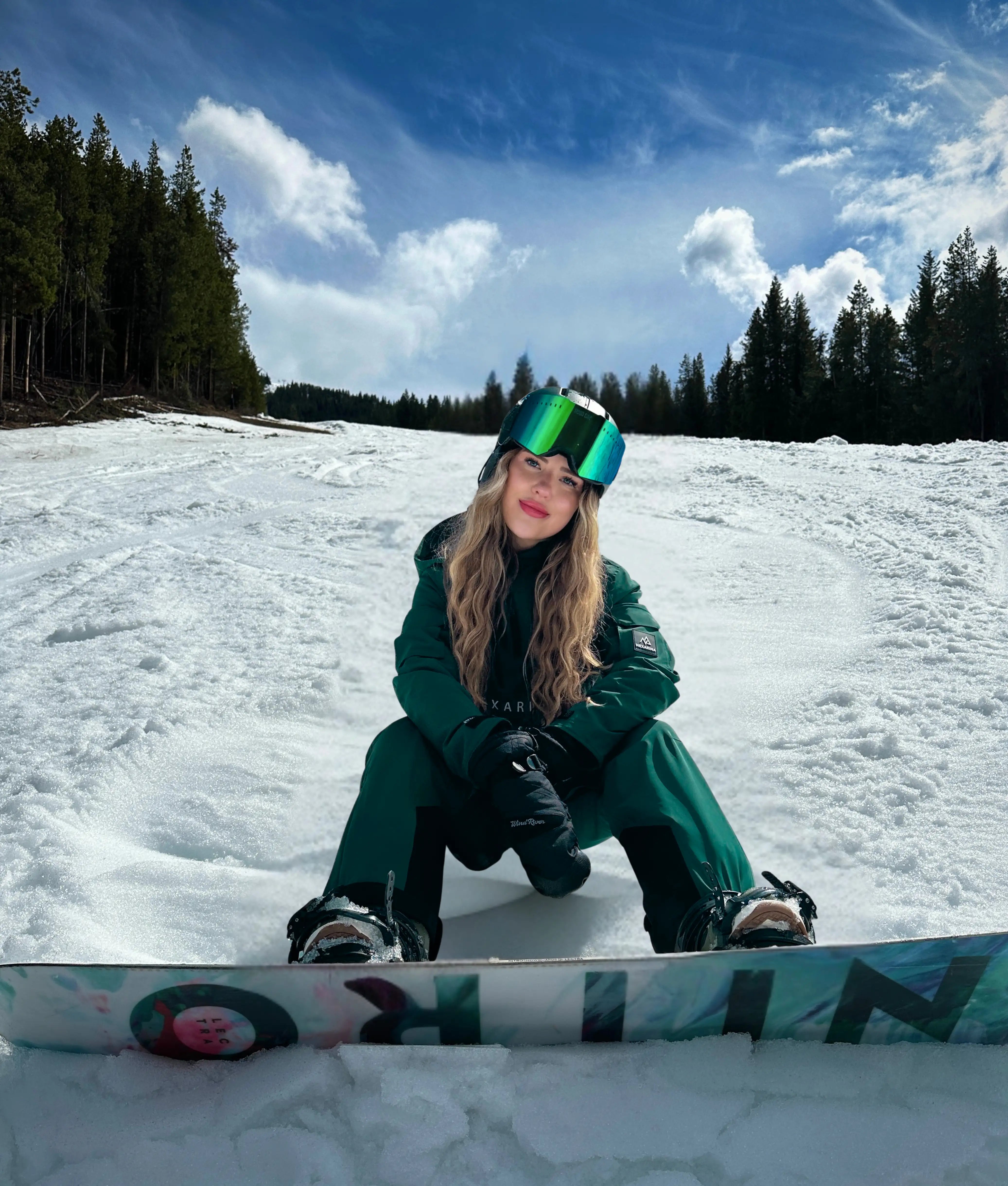 Female snowboarder wearing Nexarina green bib pants in a mountain snow setting.