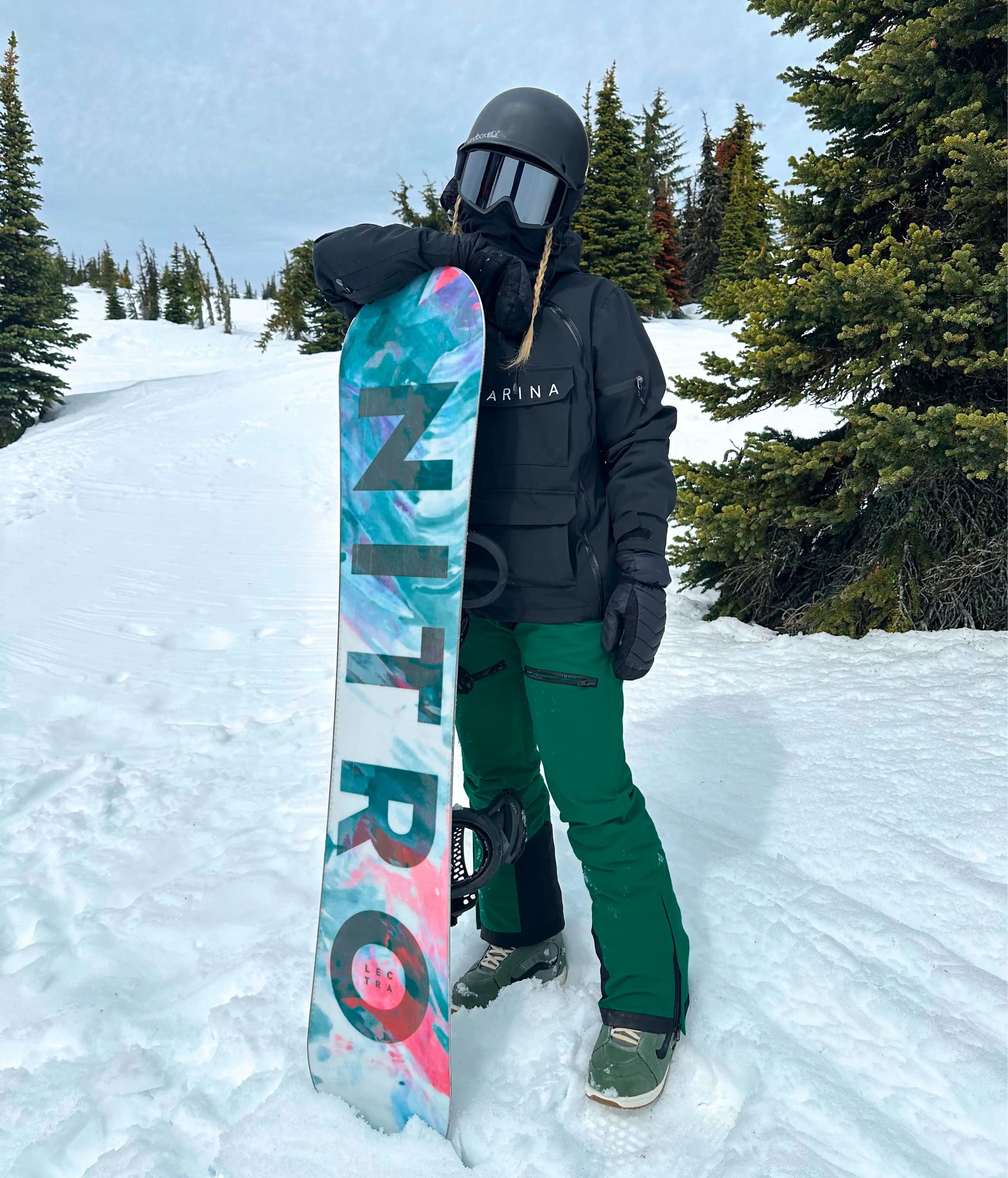 Woman standing in snowy mountain terrain holding a colorful Nitro snowboard, wearing a black Nexarina jacket and emerald green snow pants, helmet and goggles on, representing women’s technical outerwear.