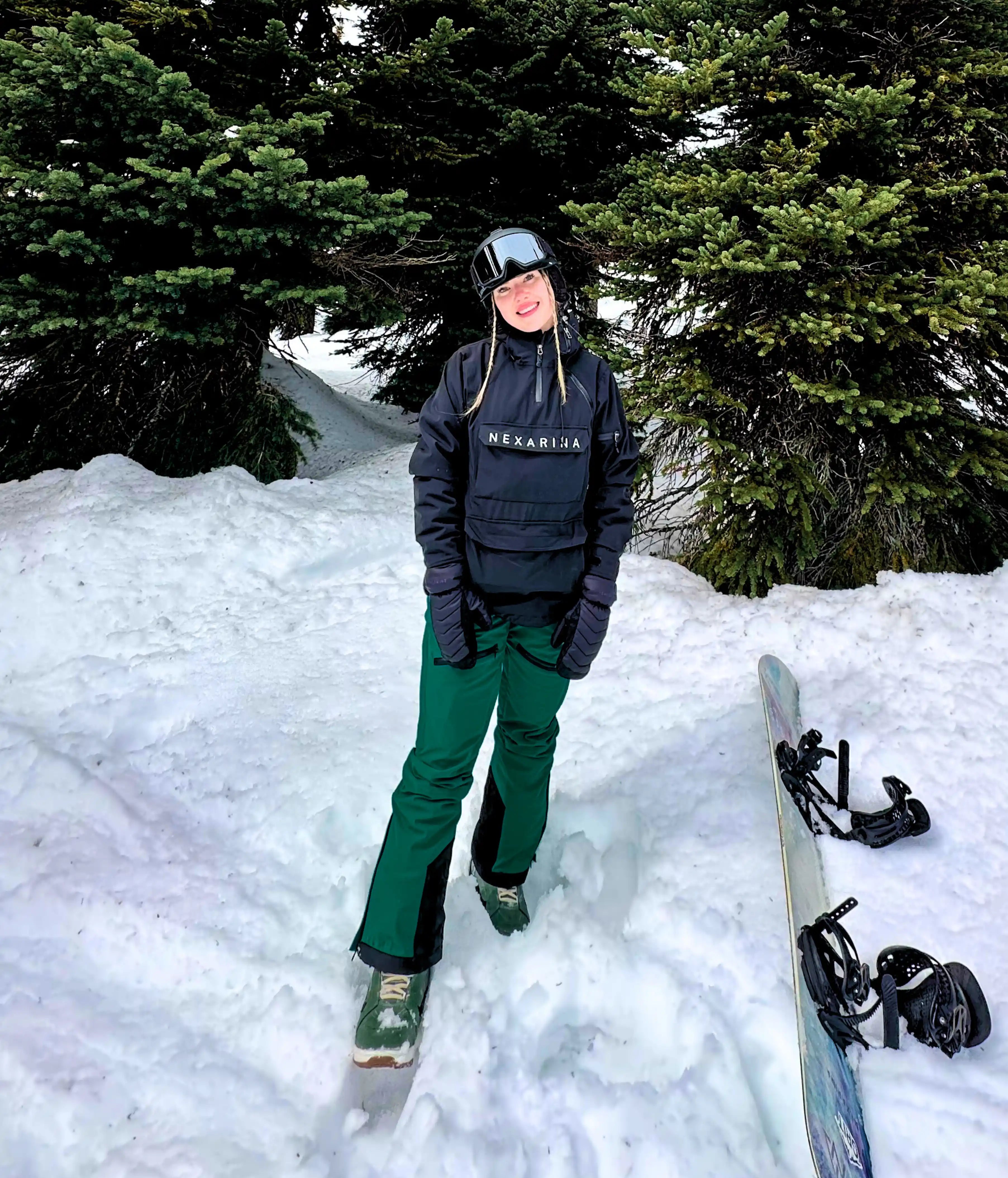 Woman smiling in snowy forest setting, dressed in a black Nexarina jacket and emerald green snow pants, helmet and goggles off, with a snowboard resting in the snow beside her.
