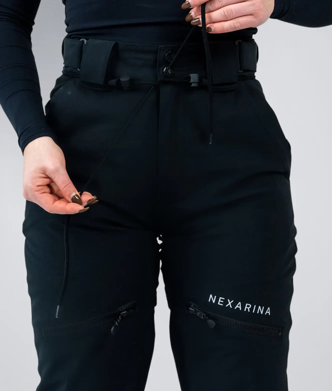 Front view of Nexarina Avani Snow Pant in Classic Black, featuring zippered thigh pockets and green boots.