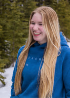 Women's Everly Hoodie in Dusty Blue worn outdoors in falling snow with the Nexarina logo visible.