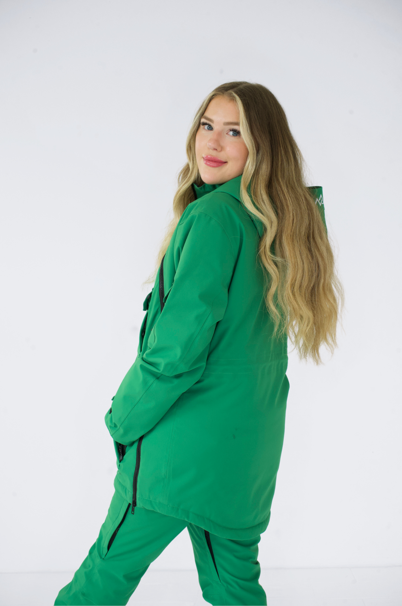 Model looking back while wearing a green Nexarina Indy snow jacket, showing the back and hood of the jacket.