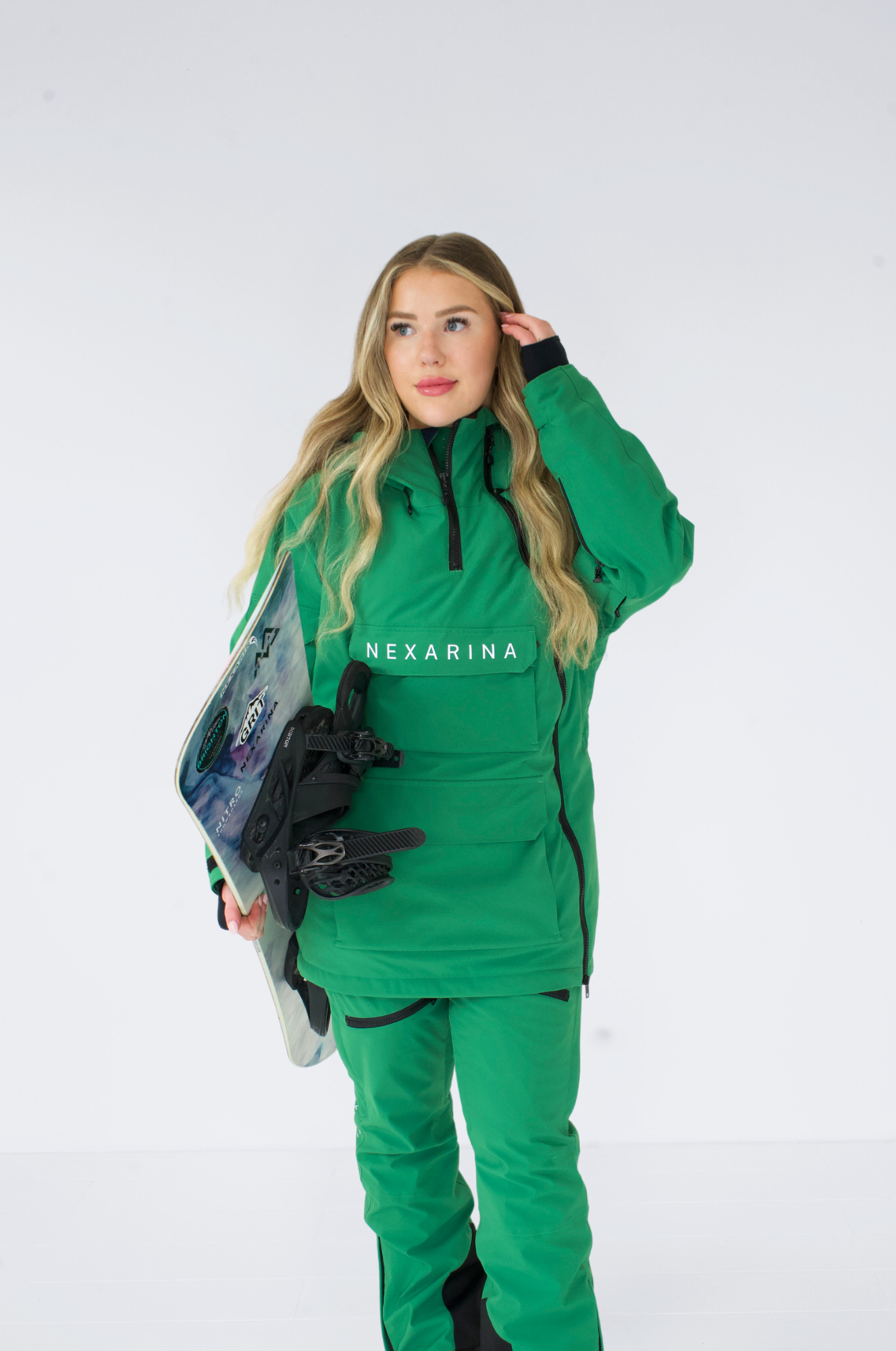 Model wearing a green Nexarina Indy snow jacket and holding a snowboard, ready for action.